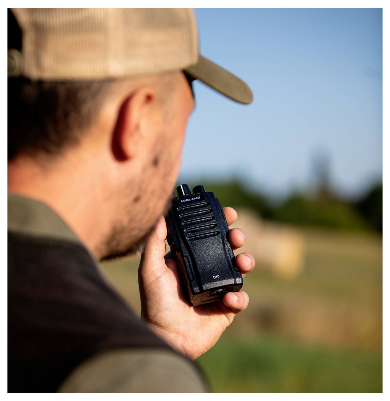 A person in a hat holds a walkie-talkie outdoors, suggesting communication in a rural or natural setting.