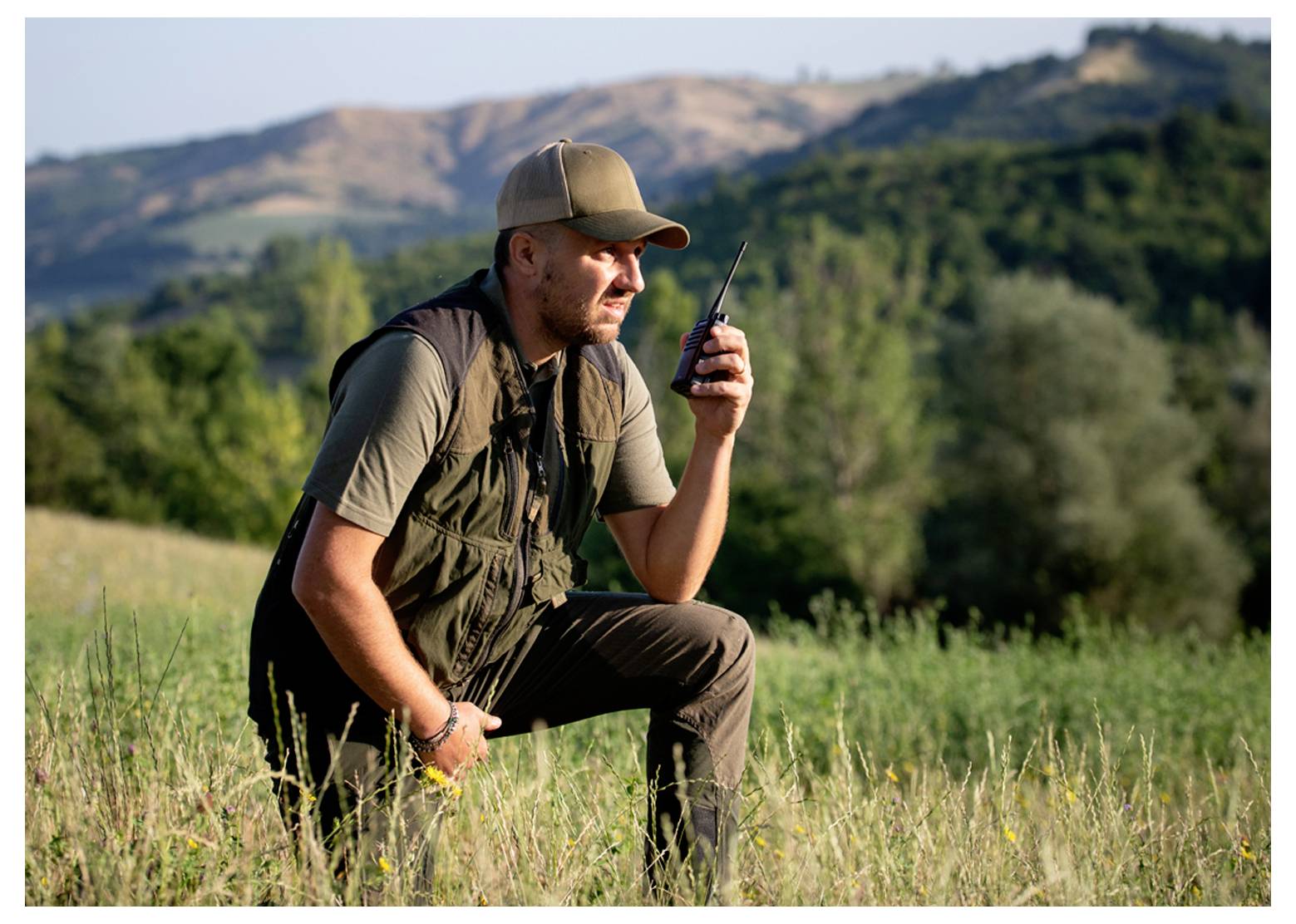 A person kneels in a grassy field, holding a walkie-talkie, with rolling hills and trees in the background.