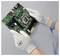 'Person holding a green circuit board with electronic components, wearing protective gloves and a wrist strap for static control.'