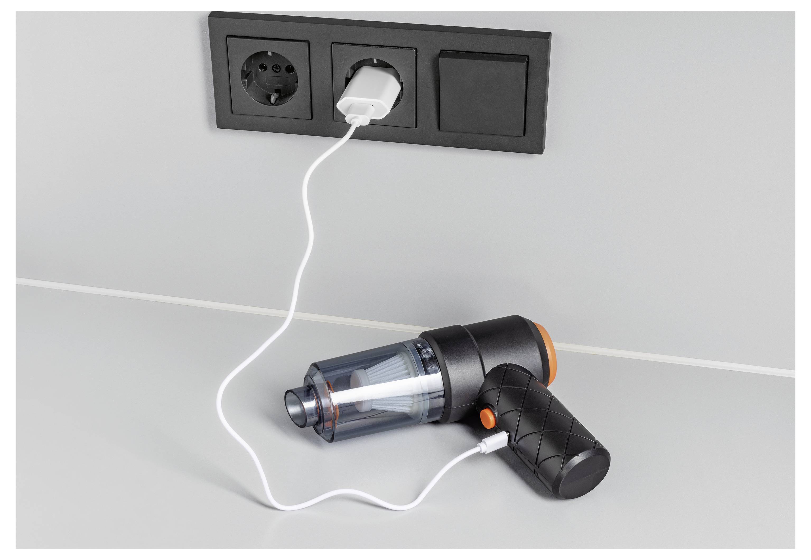 A handheld vacuum cleaner is charging on a white surface, plugged into a wall socket with a USB adapter.