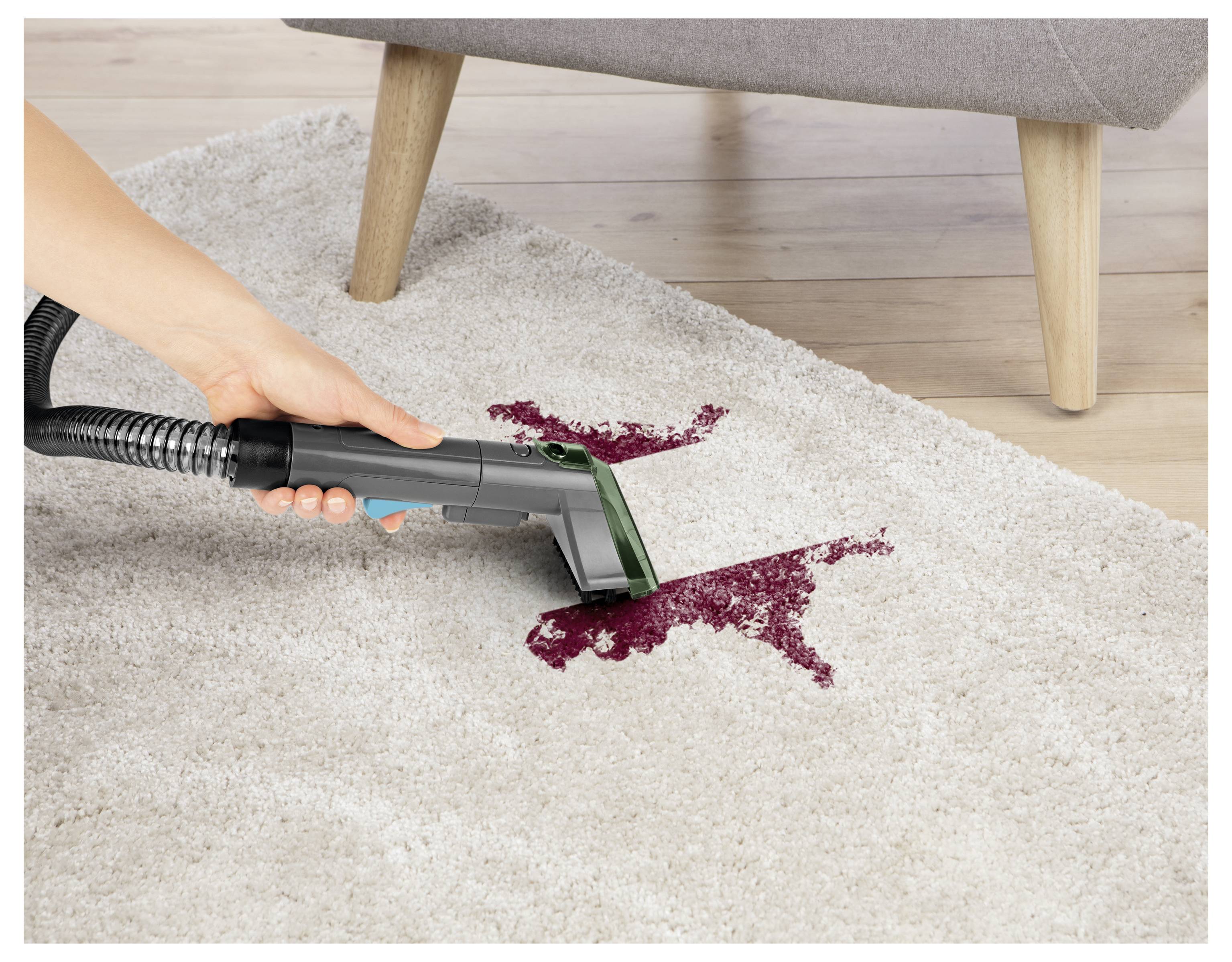 A person uses a vacuum cleaner hose to remove a red liquid stain from a white carpet, with part of a gray sofa visible in the background.