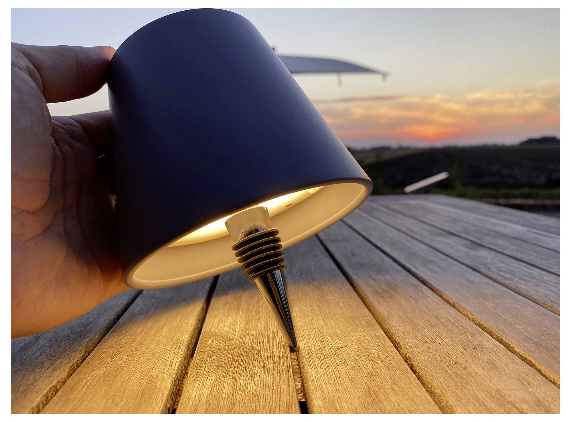 A hand holds a small, black outdoor light fixture with a spike, positioned on a wooden table during sunset, highlighting its design.