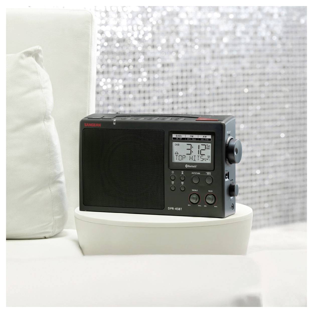 A portable radio with a digital display showing '3:12.' It is placed on a white surface next to a white pillow and a sparkling background.