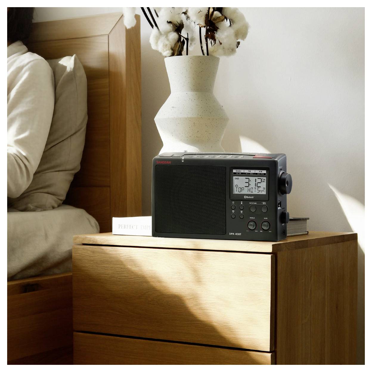A digital radio displaying the time '3:12 PM' sits on a wooden nightstand, with decorative pottery and cotton stems.
