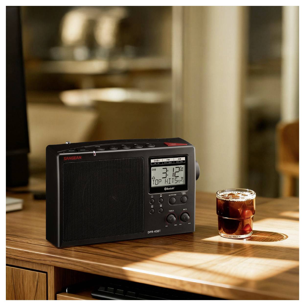 A digital radio with a display showing '3:12' and '101.5 MHz' is on a wooden table beside a glass of dark beverage.
