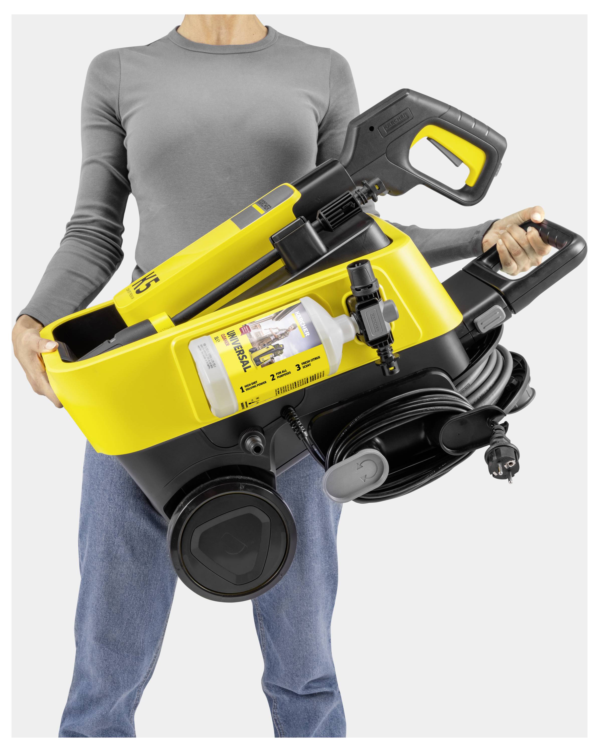 A person holds a portable pressure washer with a spray gun, yellow body, and black handle. Coiled cable and detergent bottle included.