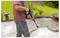 'Person using a pressure washer to clean a concrete patio, with garden plants in the background on a sunny day.'
