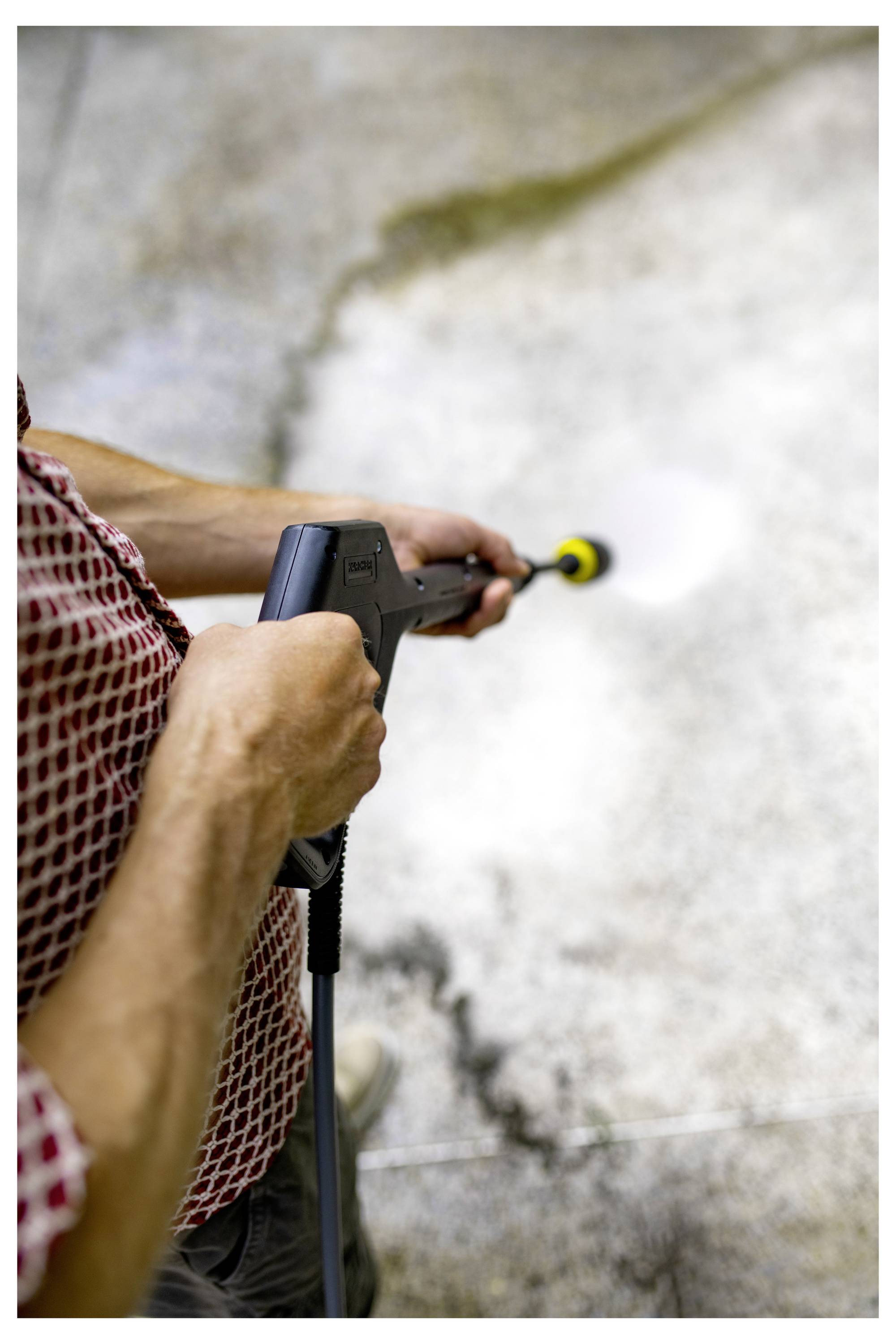 A person in a red checkered shirt is using a pressure washer to clean a concrete surface, with a clear contrast between clean and dirty areas.