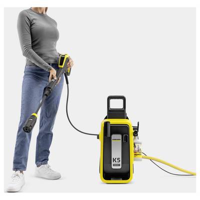 A person using a Kärcher K5 pressure washer connected by hose, holding the trigger gun, dressed in casual clothing, set against a neutral background.