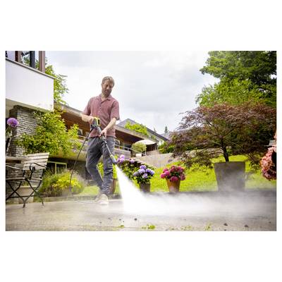 A person in a garden using a high-pressure washer to clean a patio. Flowers and trees surround the area, creating a serene setting.