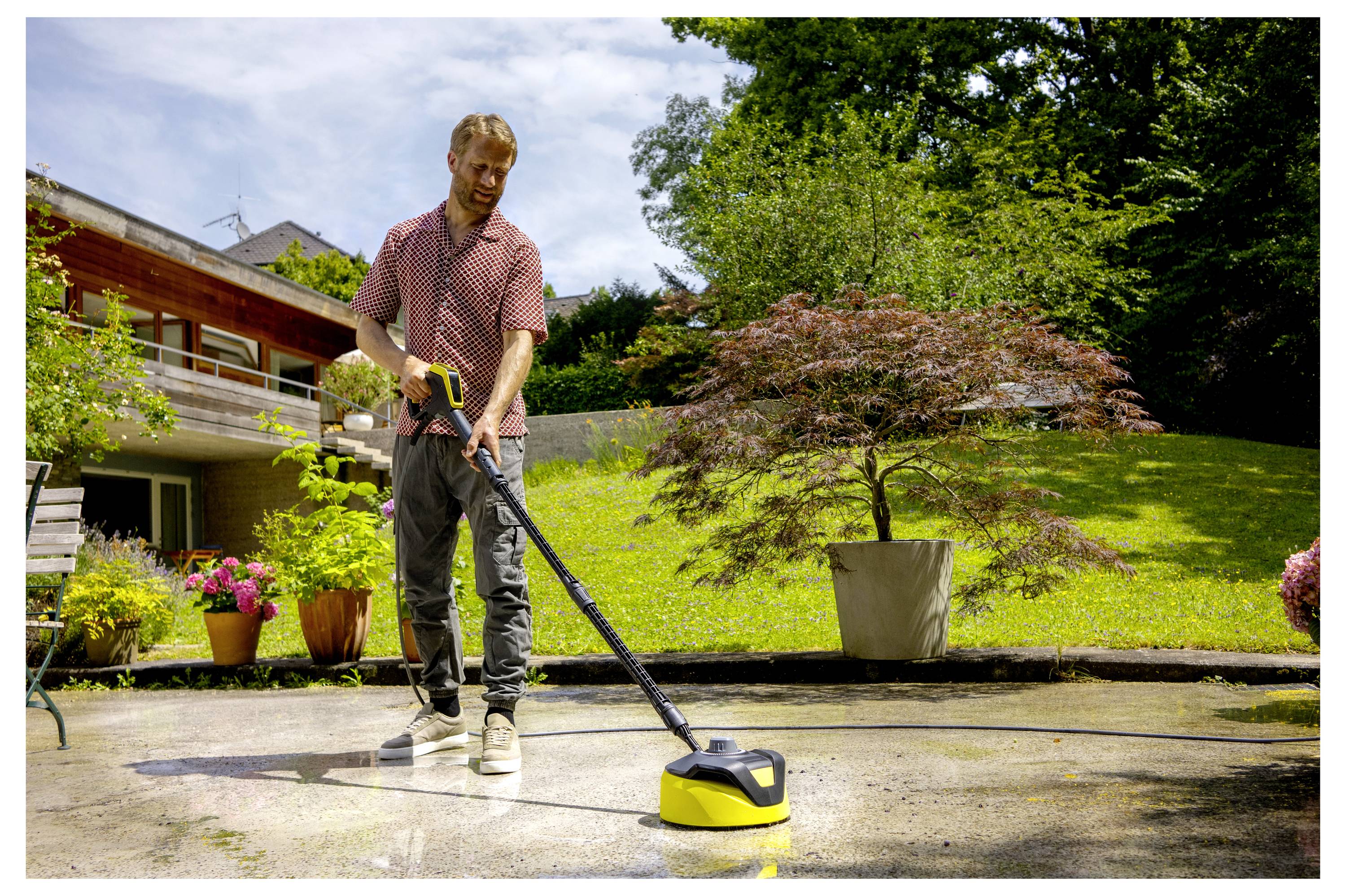 A person uses a pressure washer to clean a patio in a garden with potted plants and a large tree in the background.