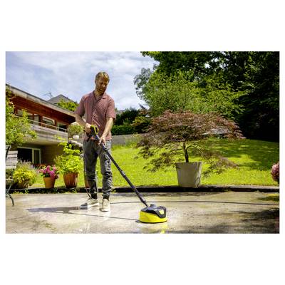 A person uses a pressure washer to clean a patio in a garden with potted plants and a large tree in the background.
