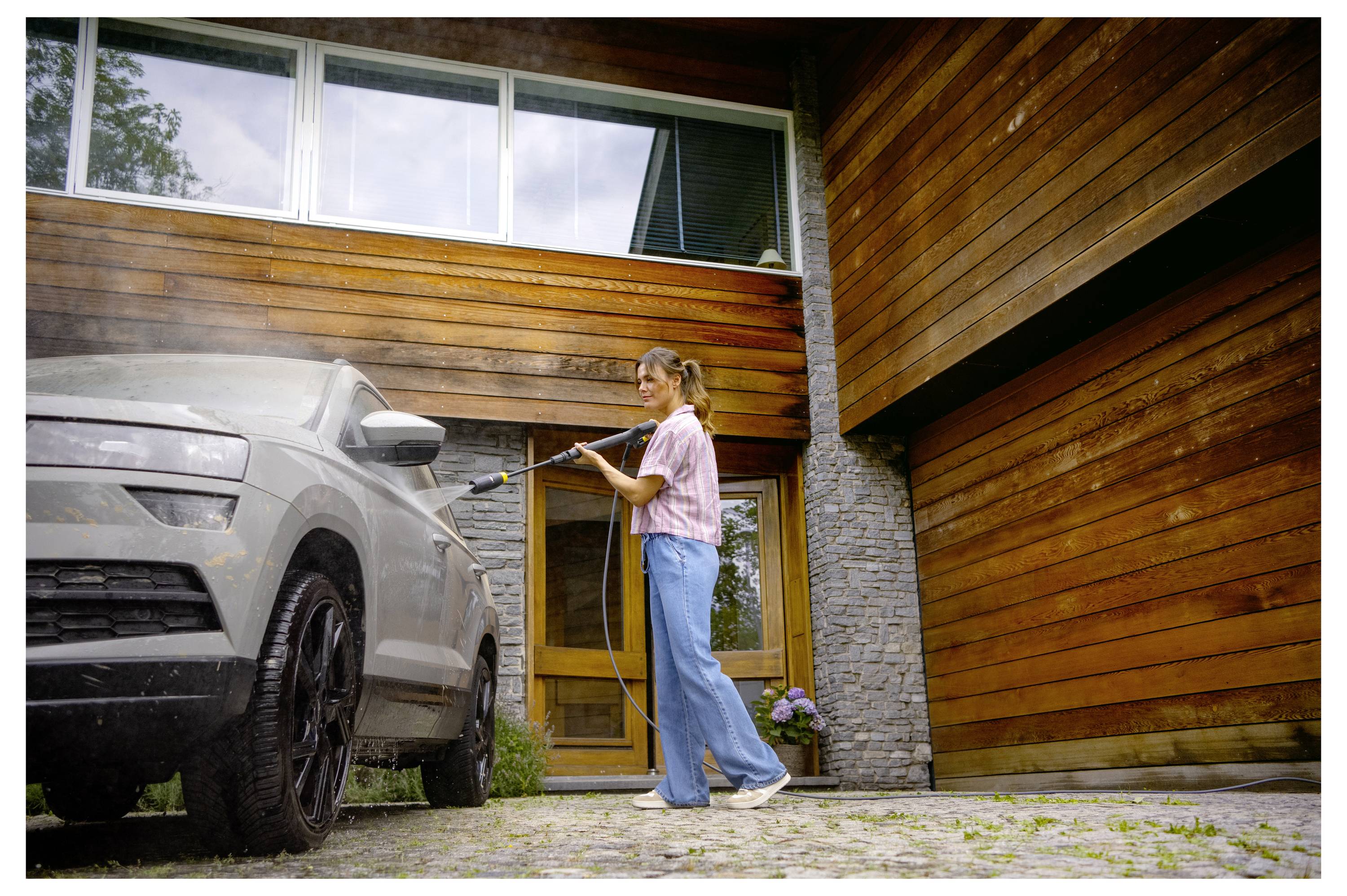 A person is pressure washing a car in front of a modern wooden house, with large windows visible in the background.