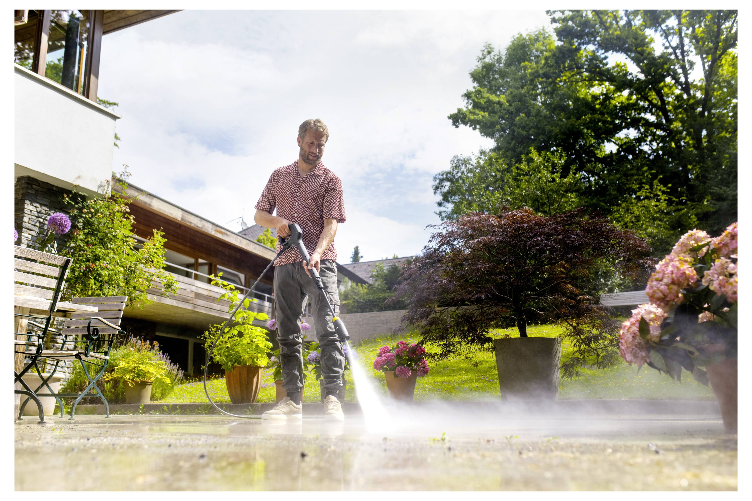 A man uses a pressure washer to clean a patio surrounded by potted plants and outdoor furniture, with a building and trees in the background.