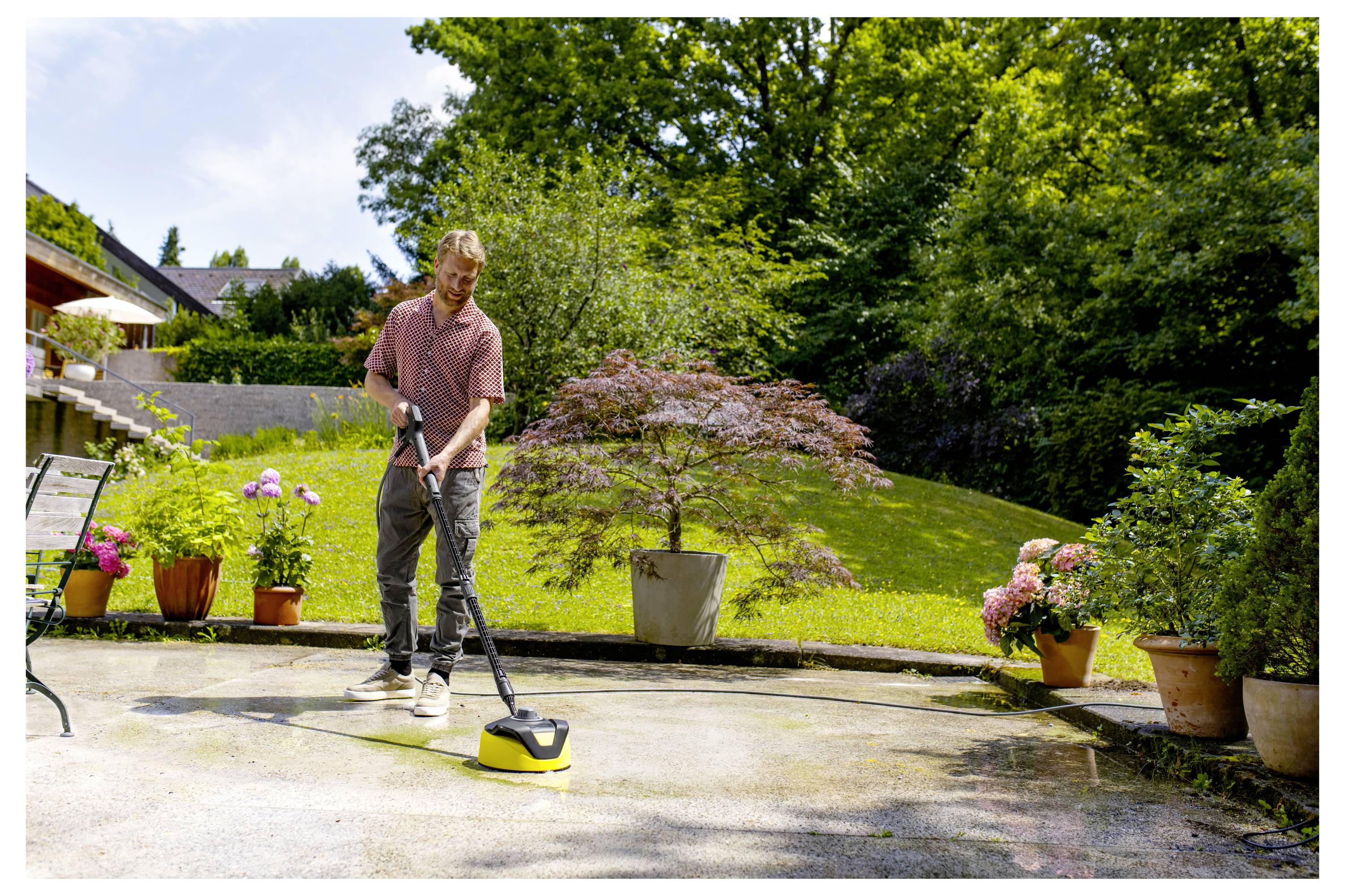 A man uses a power washer to clean a patio in a sunny garden with trees and potted plants around.