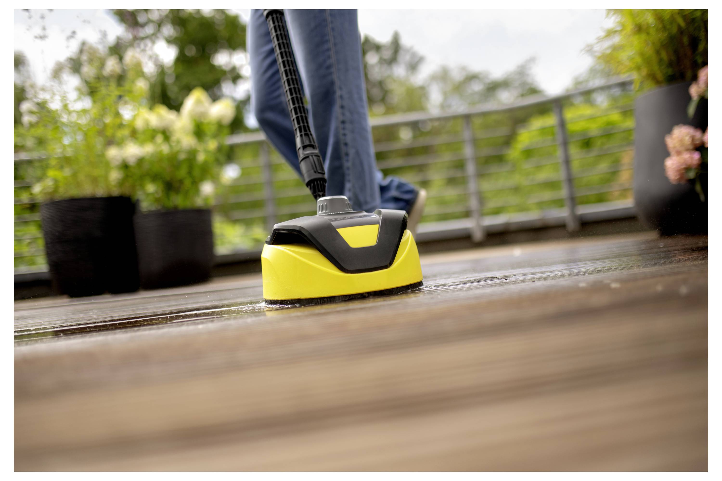 A person uses a yellow pressure washer on a wooden deck, surrounded by potted plants and a metal railing in an outdoor setting.