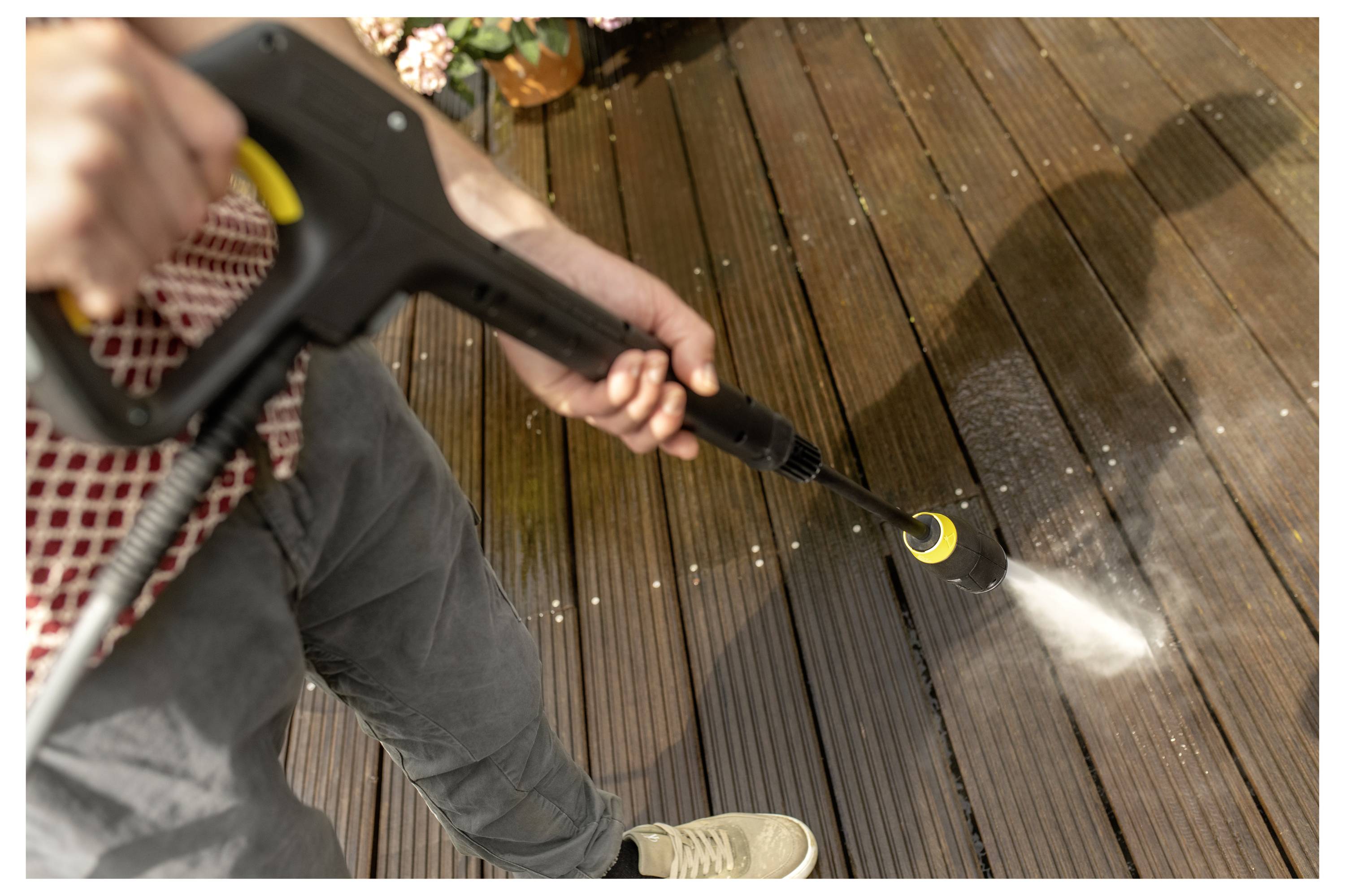 A person uses a pressure washer to clean wooden decking. Water sprays from the washer, removing dirt and leaving a clean surface.