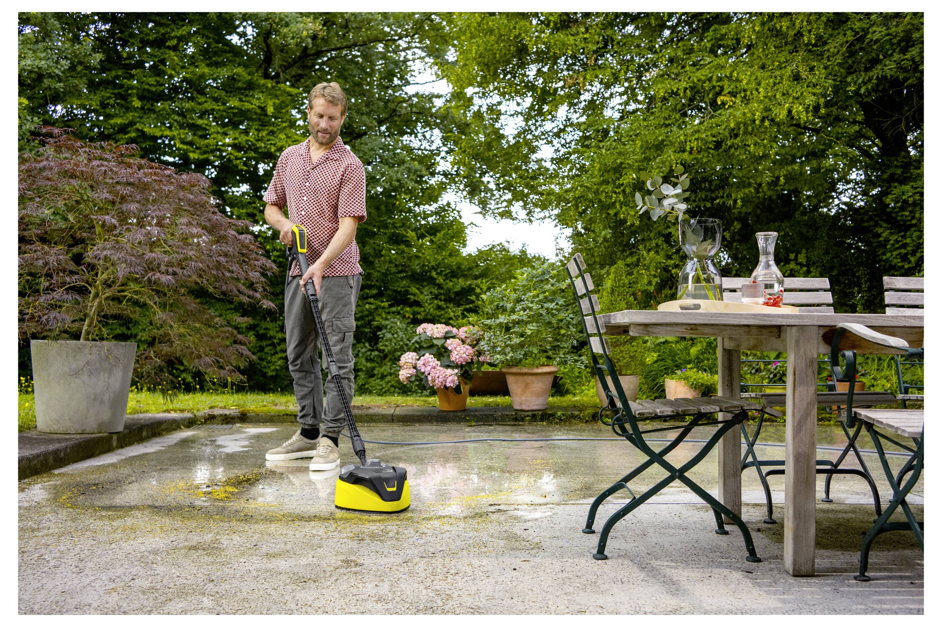 A man uses a pressure washer on an outdoor patio. A table with chairs and plants surround the area, creating a garden setting.