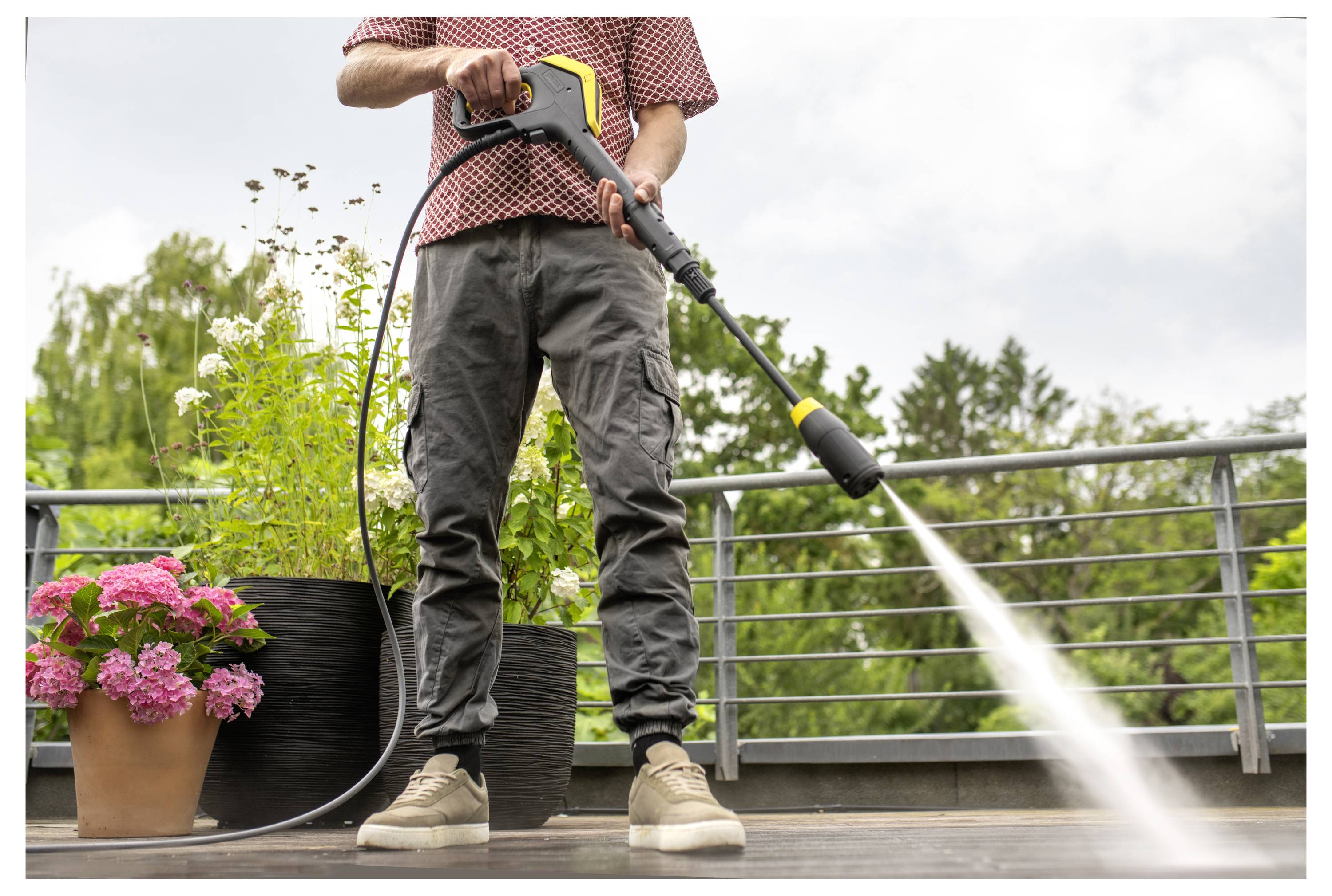 A person pressure-washes a wooden deck surrounded by potted plants, including pink and white flowers, under a cloudy sky.