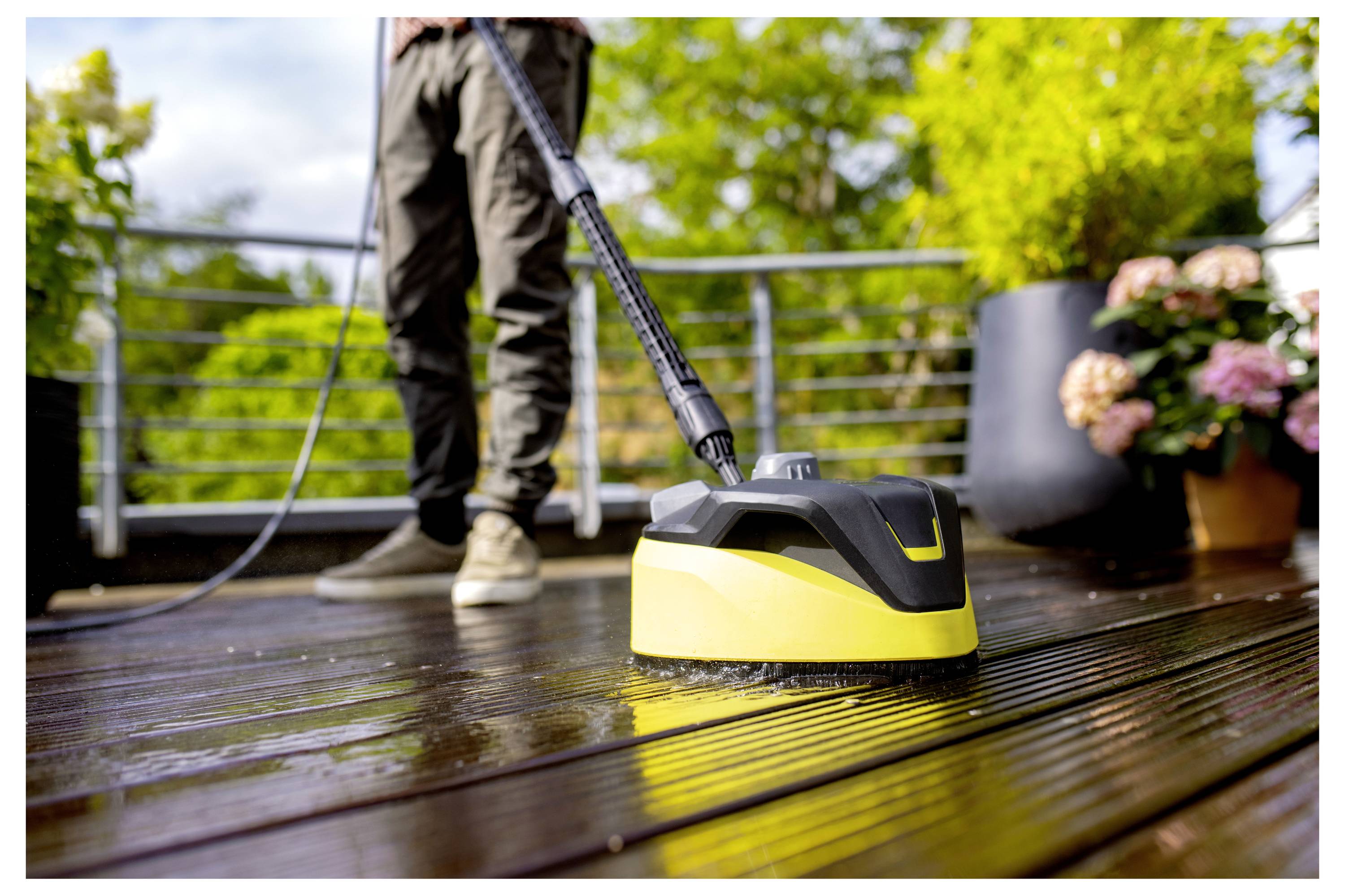 A person uses a yellow power scrubber to clean a wet wooden deck, surrounded by potted plants and greenery on a sunny day.