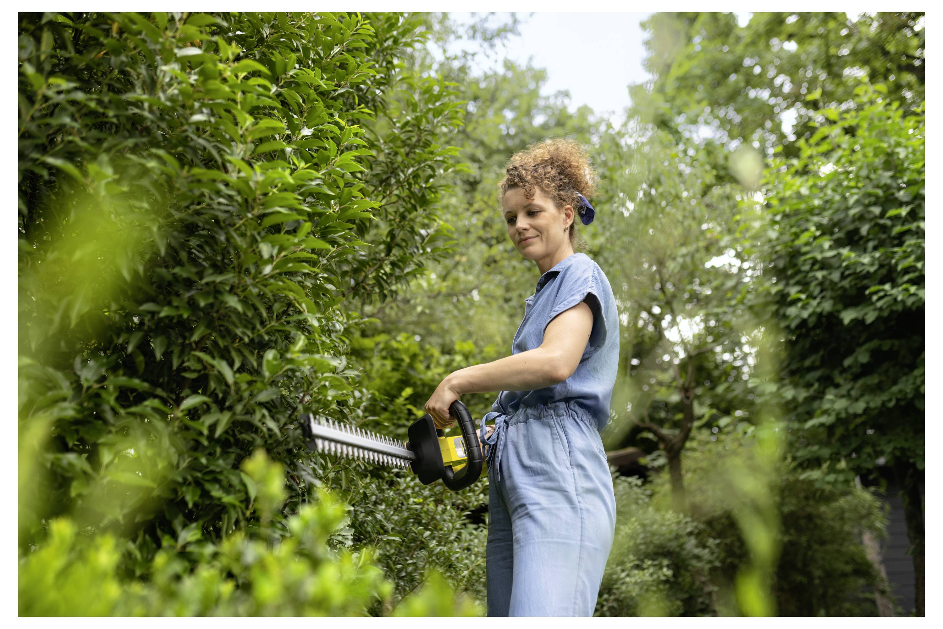 A person in a blue jumpsuit trims a lush green hedge with a power tool, surrounded by trees and greenery, in an outdoor garden setting.