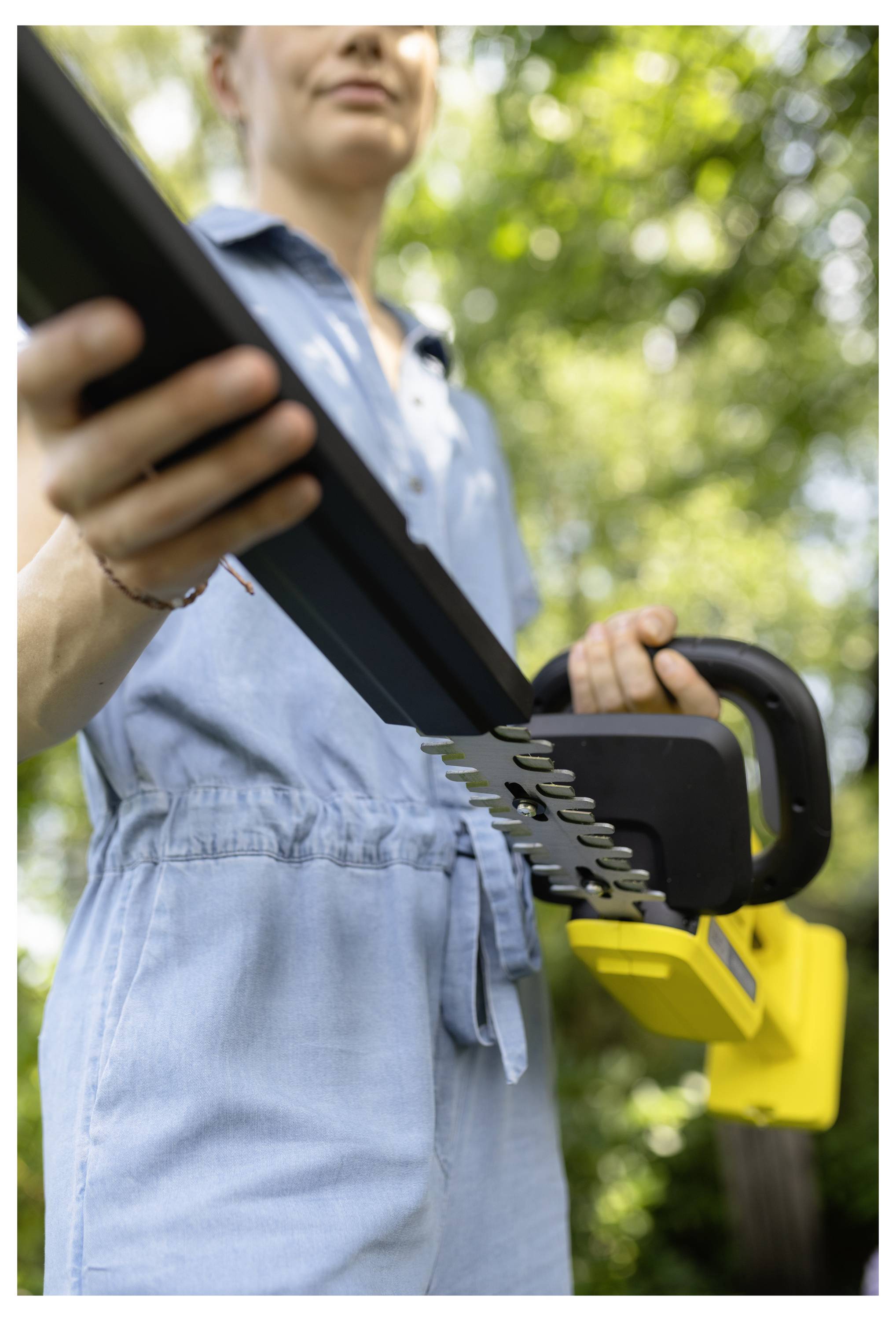 A person in a light blue jumpsuit holding a yellow electric saw outdoors, with trees in the background, suggesting a gardening task.