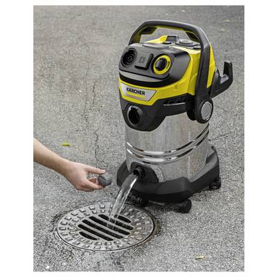 A person's hand drains water from a yellow and stainless steel wet/dry vacuum cleaner into a grate on an asphalt surface.