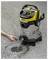 A person's hand drains water from a yellow and stainless steel wet/dry vacuum cleaner into a grate on an asphalt surface.