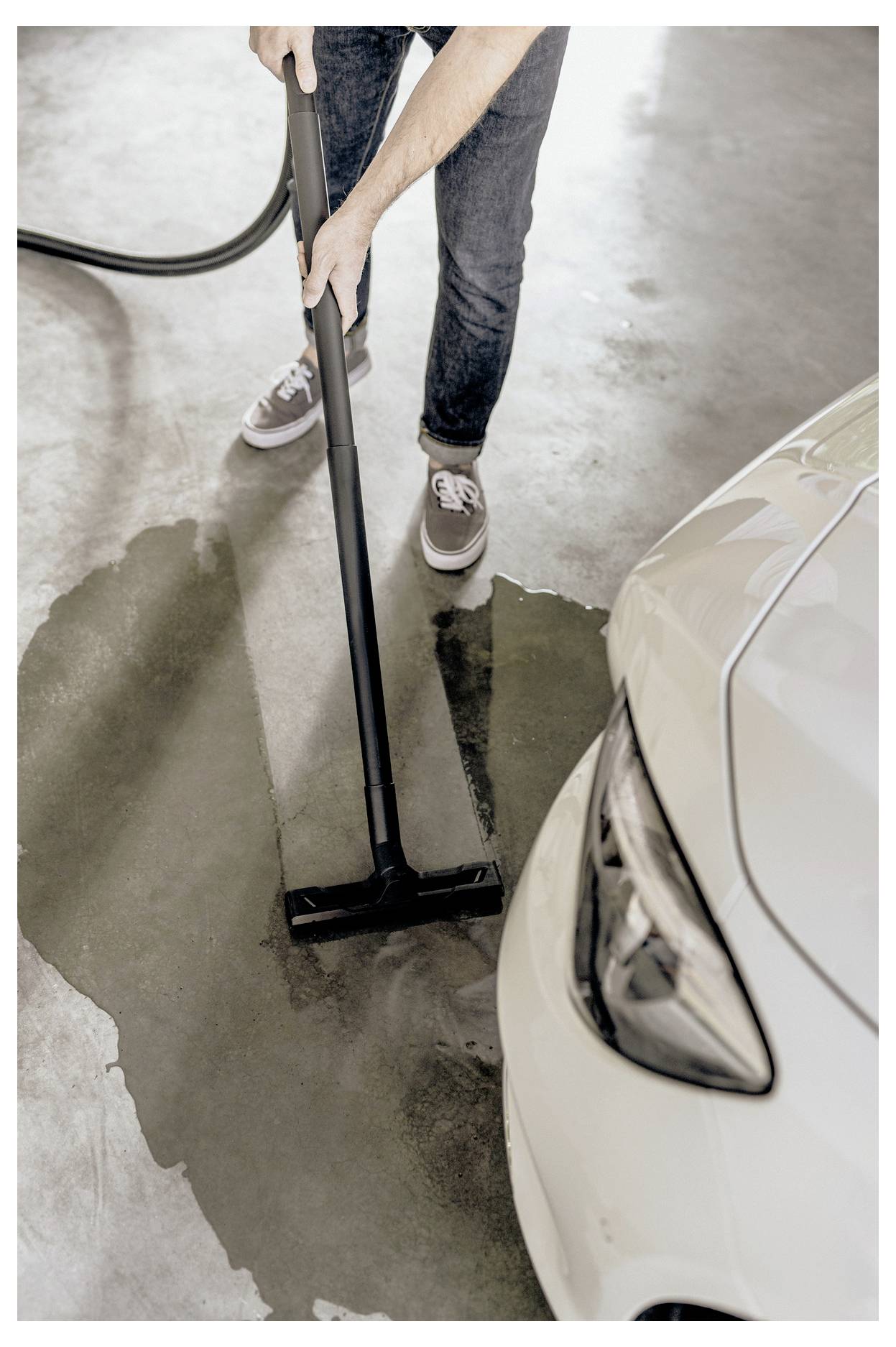 A person vacuums water from a concrete garage floor near a white car. Their hands and the vacuum handle are visible.