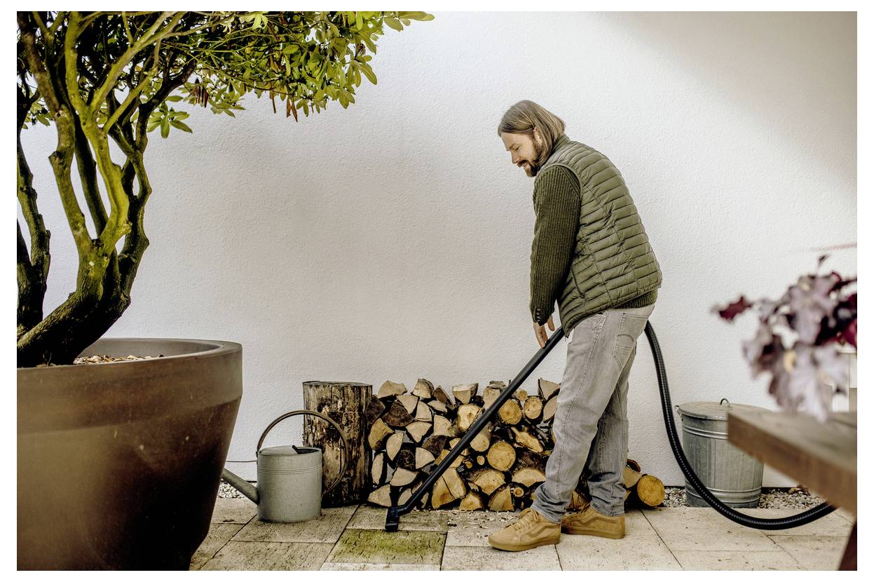 A person in a garden stacks firewood beside a tree in a large pot. Nearby, there's a watering can and a hose on a wooden patio.