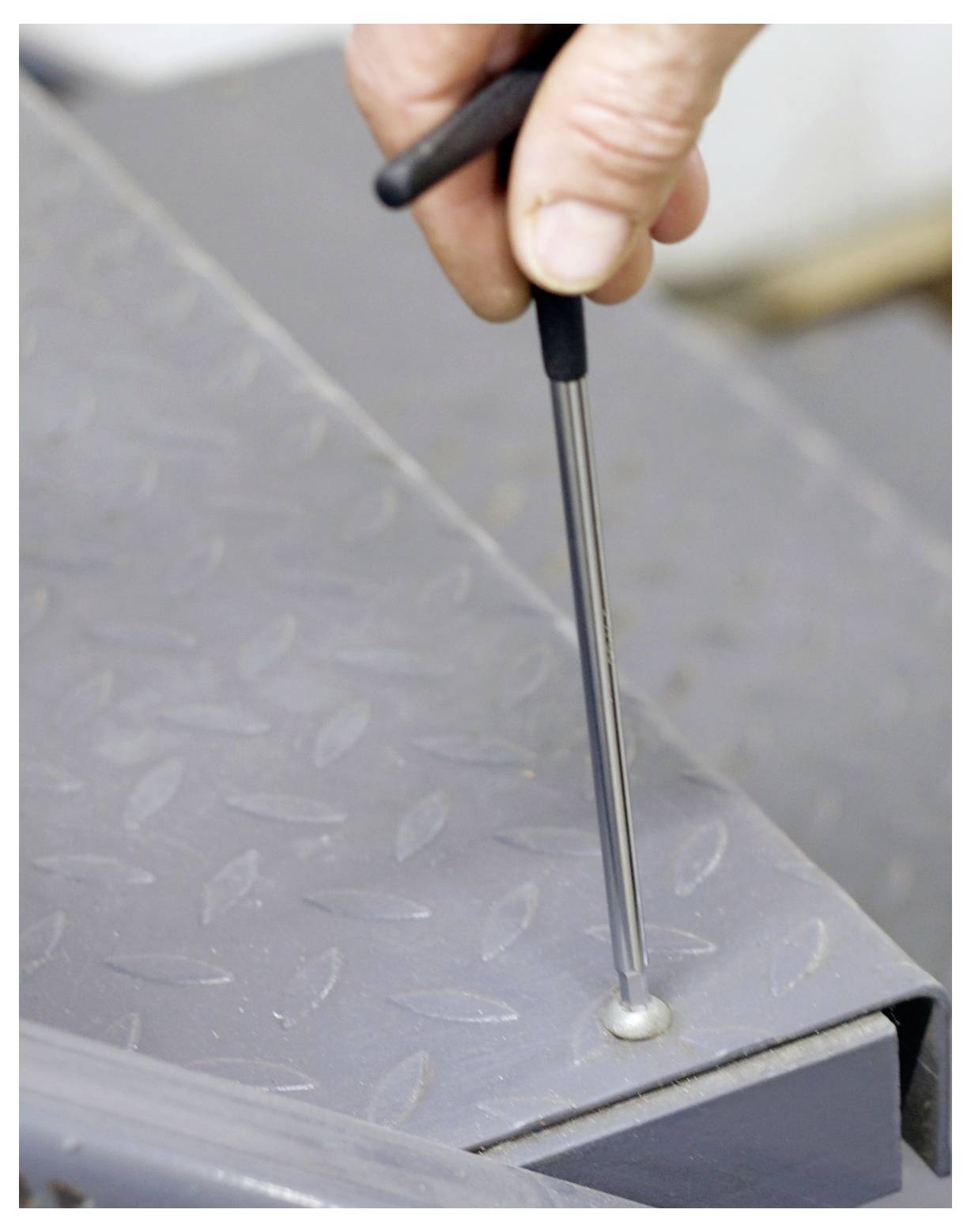 A hand uses a metal detector rod on a diamond plate surface to detect hidden objects.
