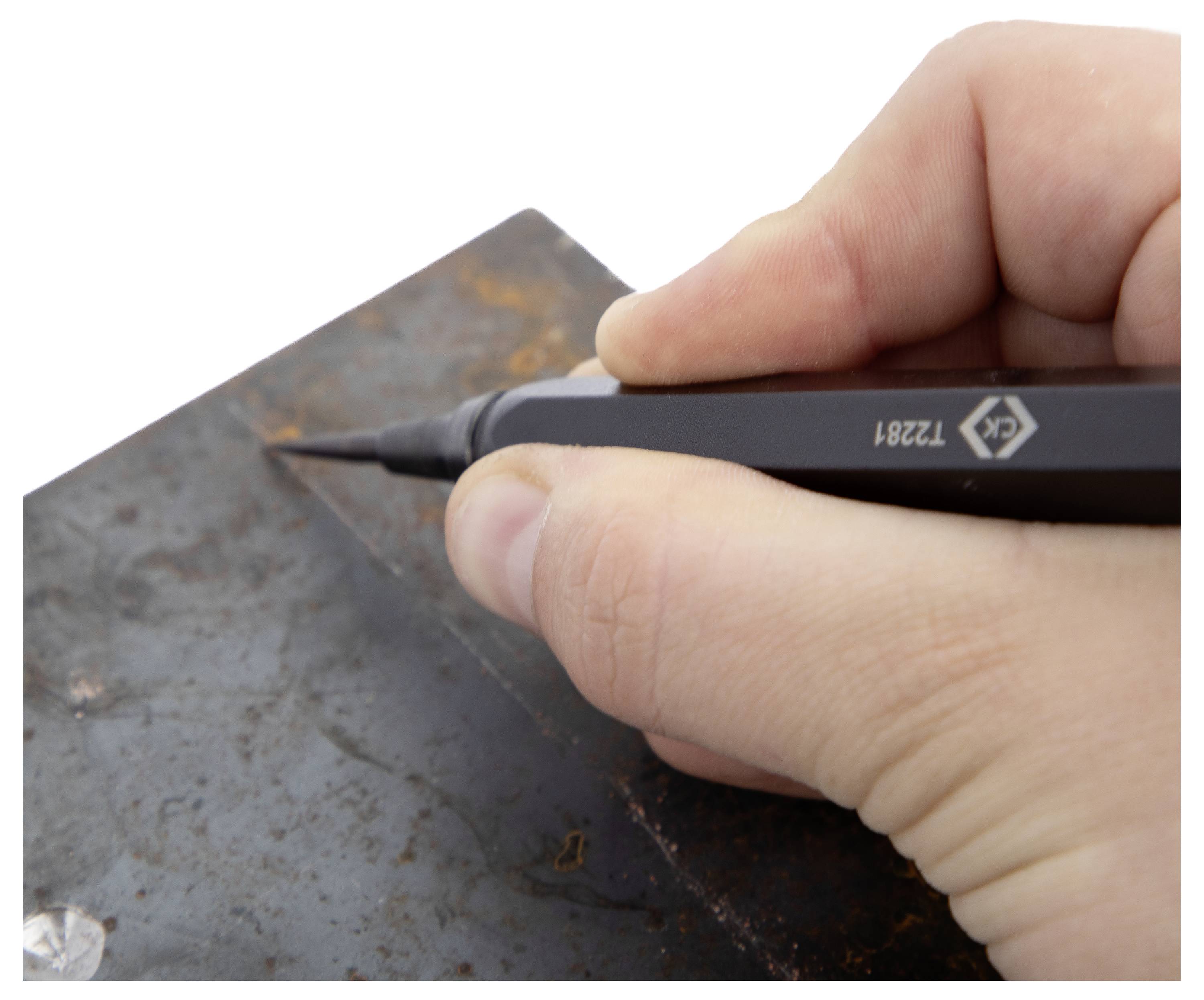 A hand holding a pen-like tool etches or marks a line on a rusted metal surface.