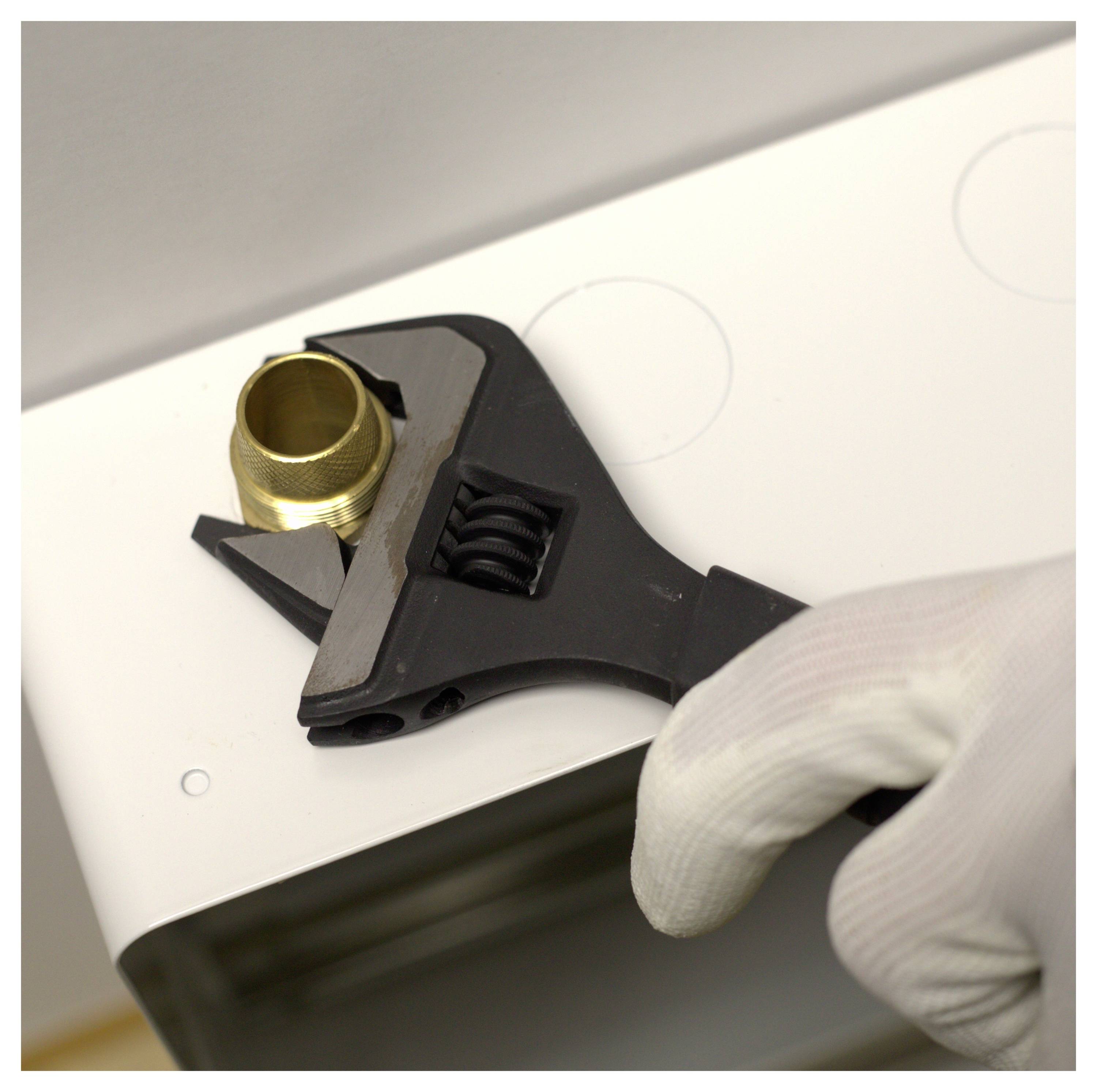 'A gloved hand uses an adjustable wrench to tighten a brass fitting on a white surface, possibly relating to plumbing or assembly work.'