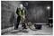 Worker in protective gear operates a jackhammer on a concrete floor in a dimly lit industrial setting.