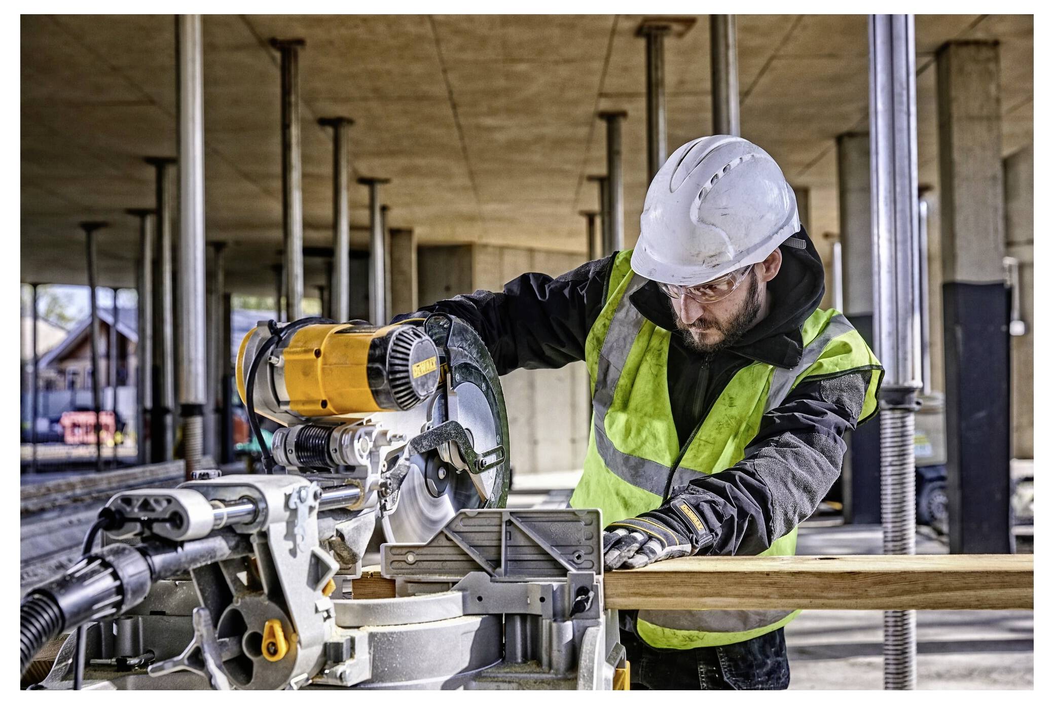'A construction worker uses a miter saw to cut a wooden board. They wear a safety helmet, high-visibility vest, and gloves on a worksite.'