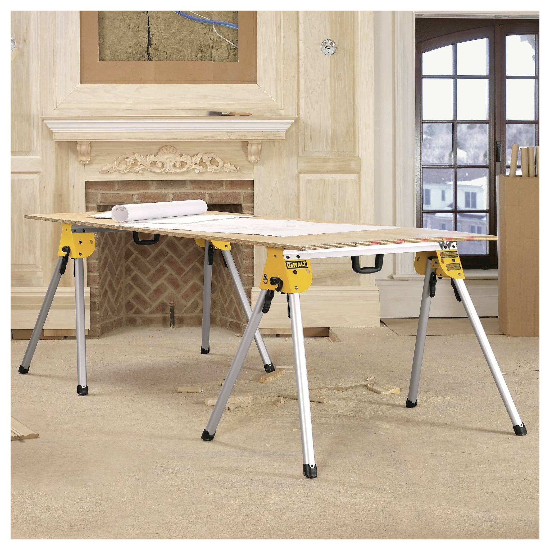 A wooden table supported by metal sawhorses in a room under renovation, with blueprints and tools nearby. Fireplace and windows in the background.