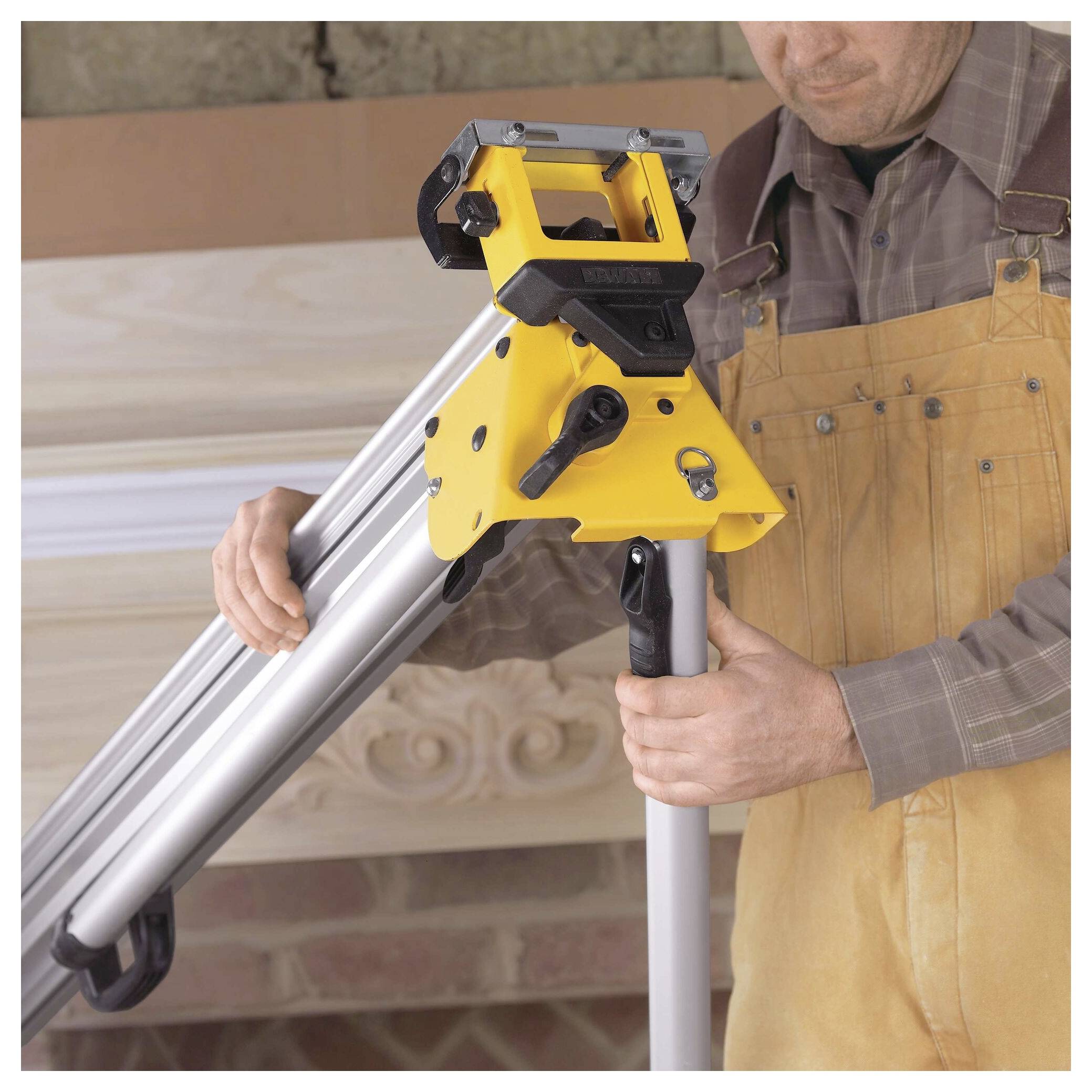 'Person in overalls adjusts a yellow saw stand with extendable metallic legs inside a workshop, surrounded by wooden planks.'