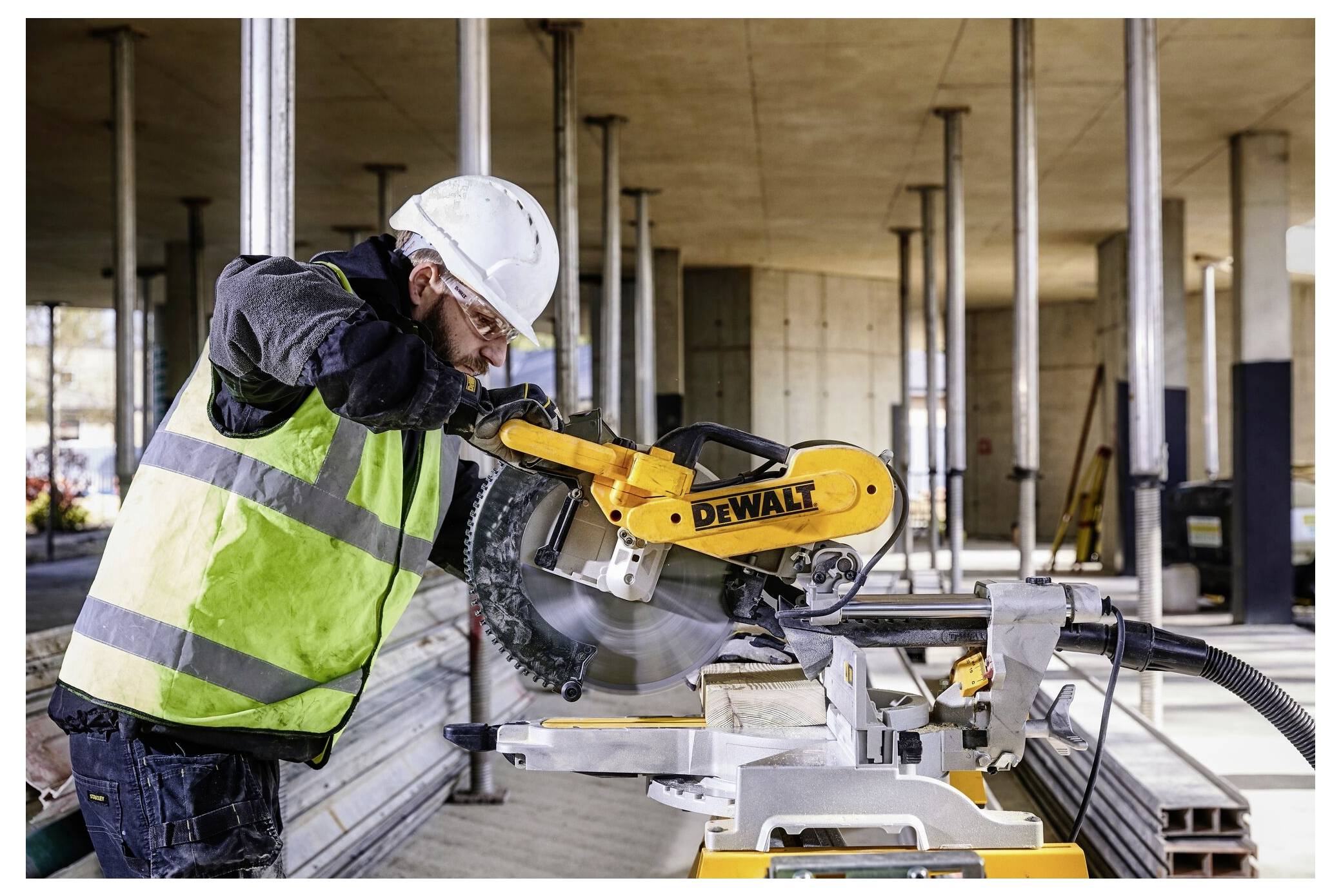 A construction worker wearing a helmet and safety vest operates a circular saw on a construction site with concrete columns and beams.