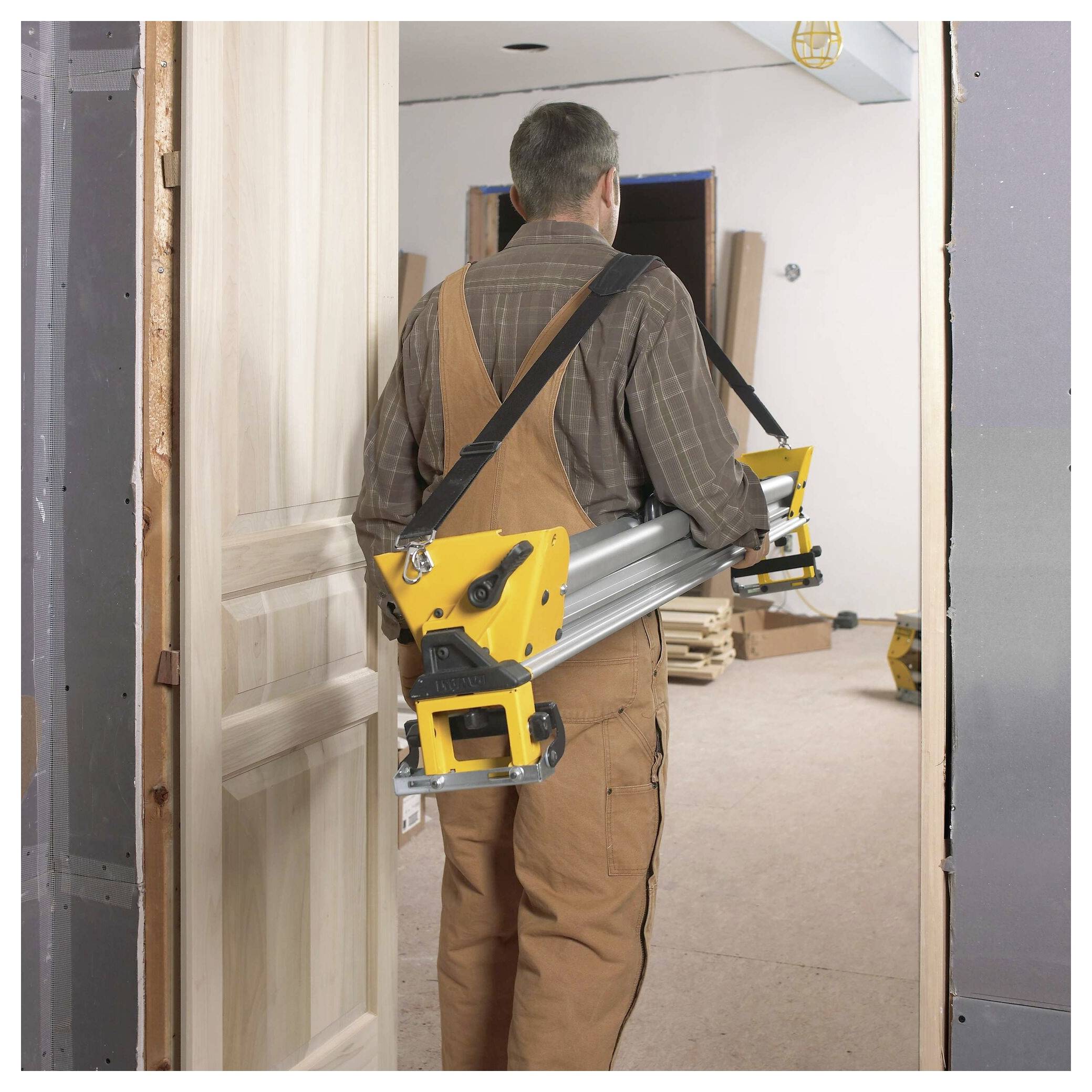 A person carrying a collapsible yellow and gray workbench through a doorway in an unfinished room, suggesting a construction or renovation setting.
