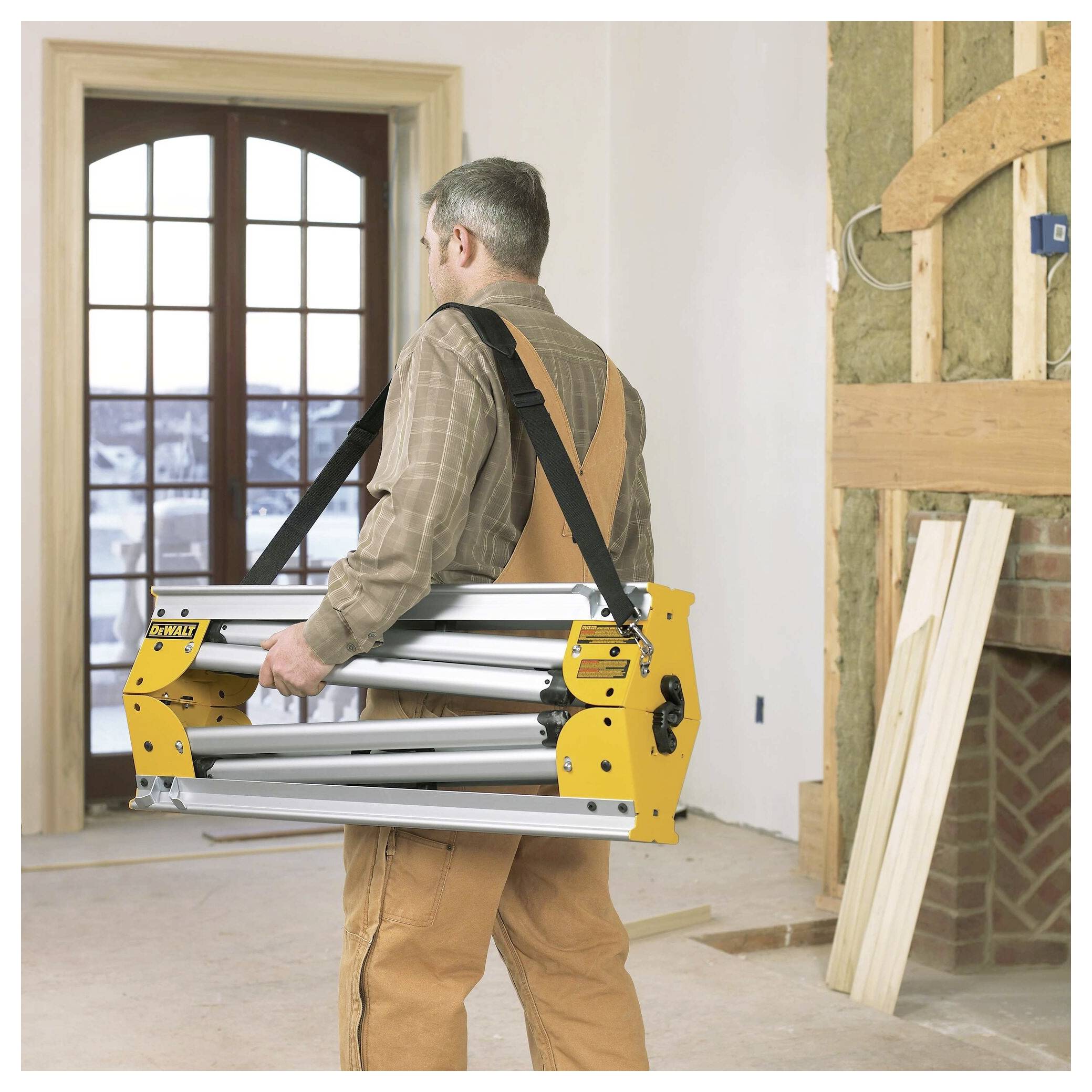 'A person in work attire carries a folded ladder inside a partially renovated room, with exposed walls and a view of snow outside.'