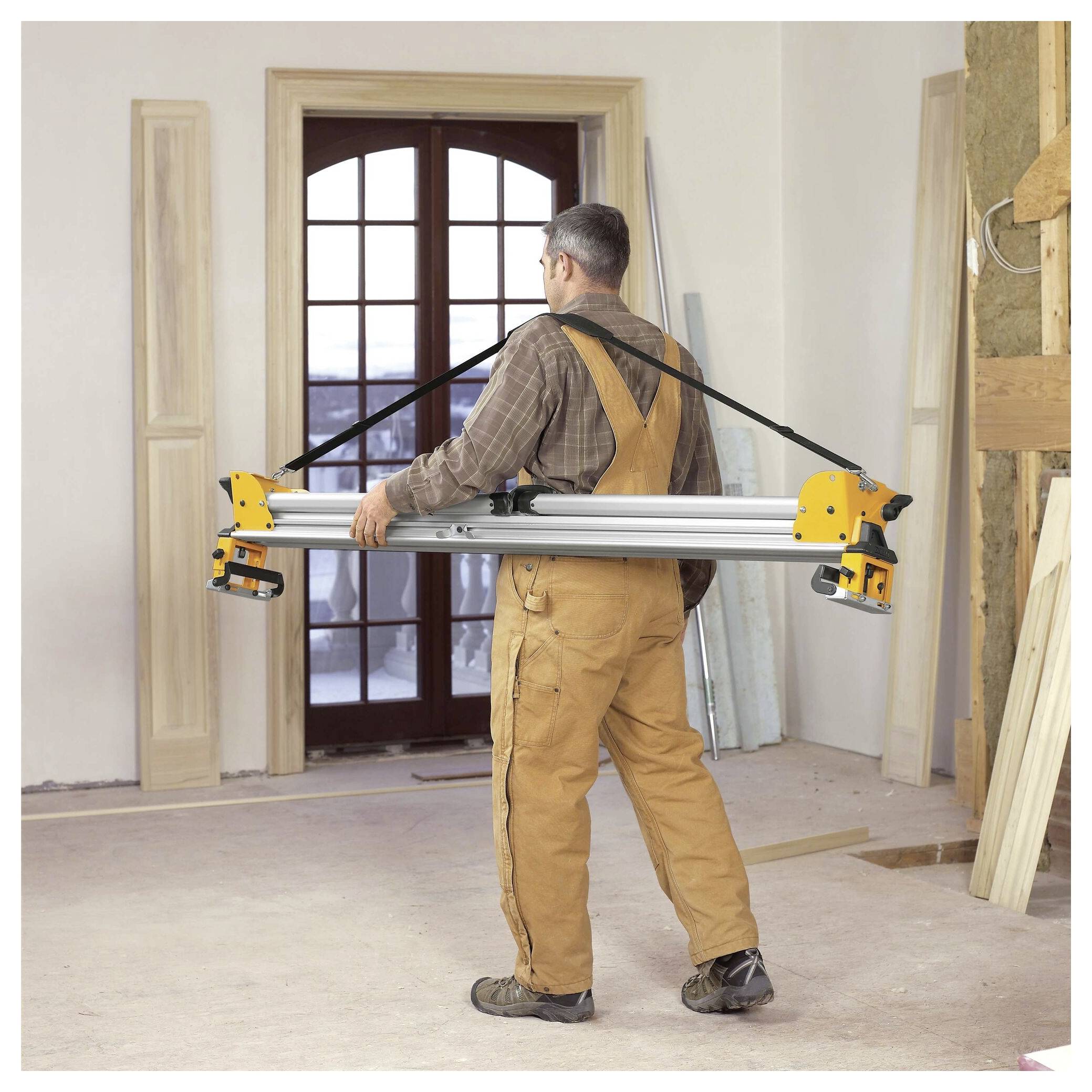 A person wearing overalls carries a large, folded sawhorse through a partially remodeled room with tools and wooden planks around.