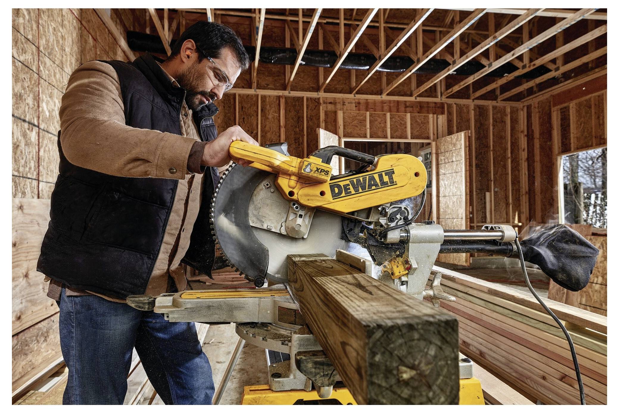 A person operates a yellow DeWalt power saw in a partially constructed wooden building, cutting through a wooden beam.