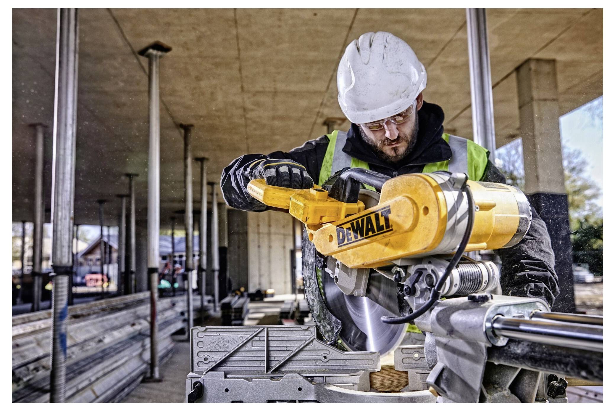 A construction worker operates a yellow power saw indoors, wearing a white helmet and high-visibility vest, focusing on cutting material.