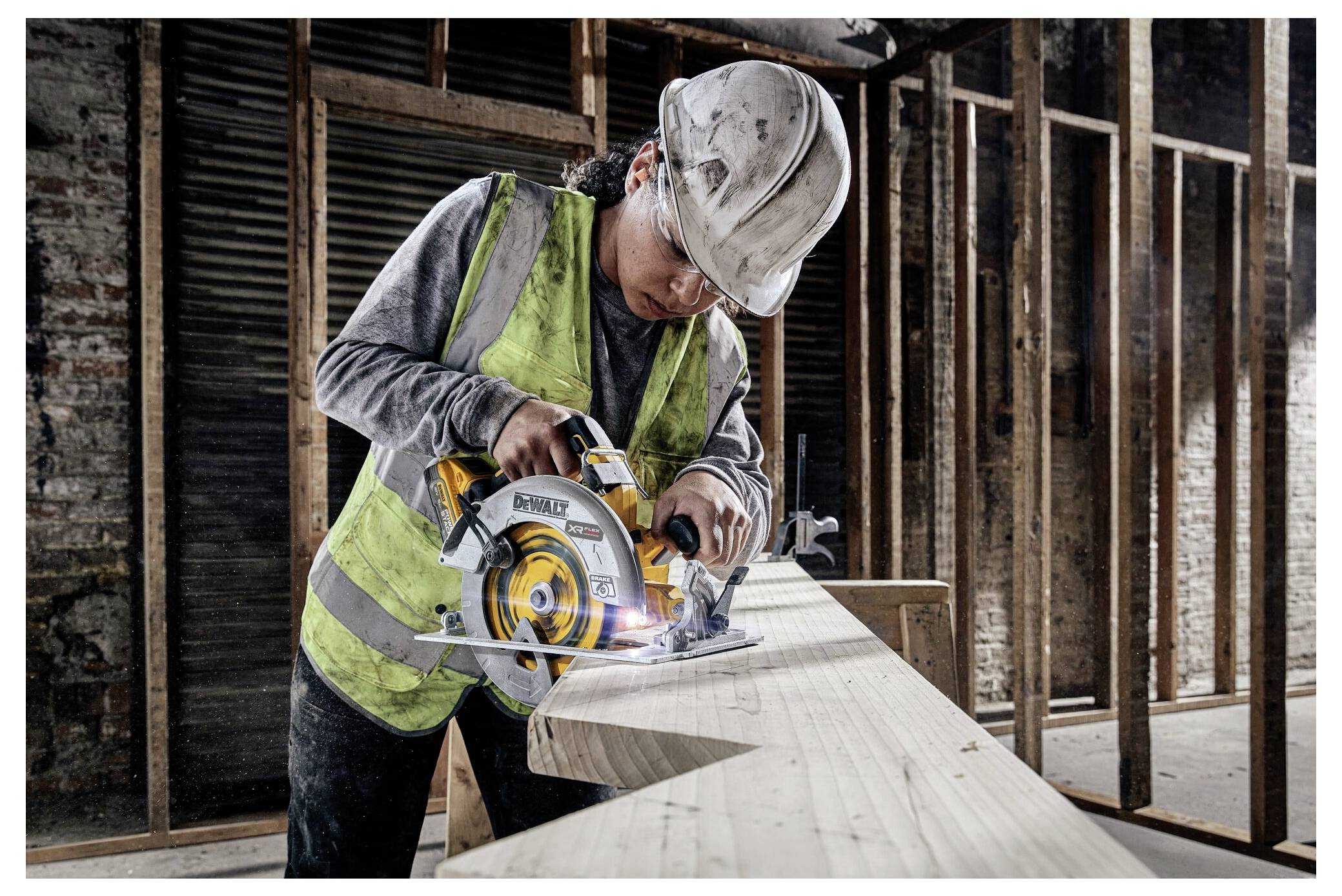 A construction worker wearing a helmet and safety vest uses a circular saw to cut a wooden beam in an unfinished building.
