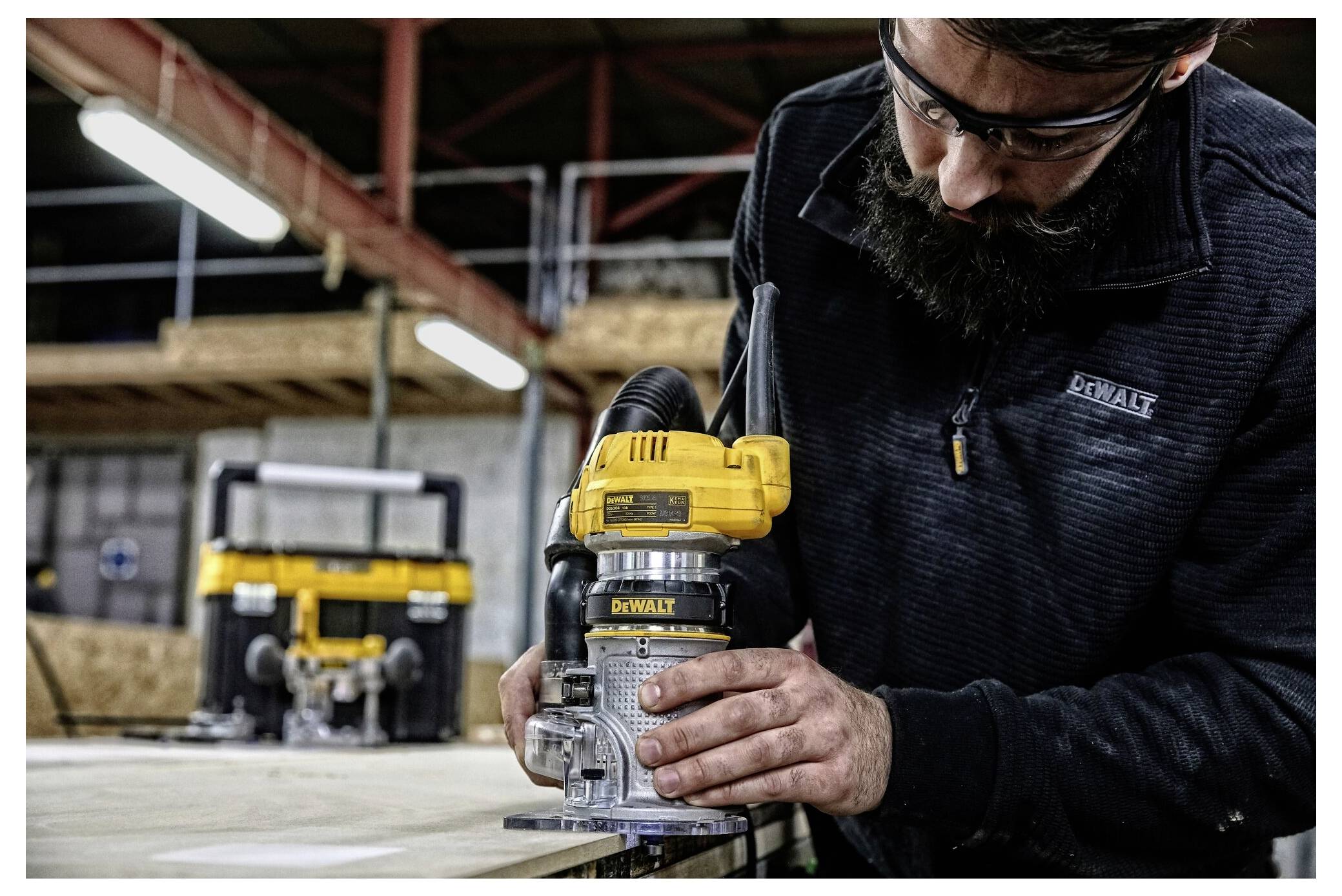 A person using a yellow and black handheld woodworking tool on a wooden surface in a workshop, focusing intently on the task.