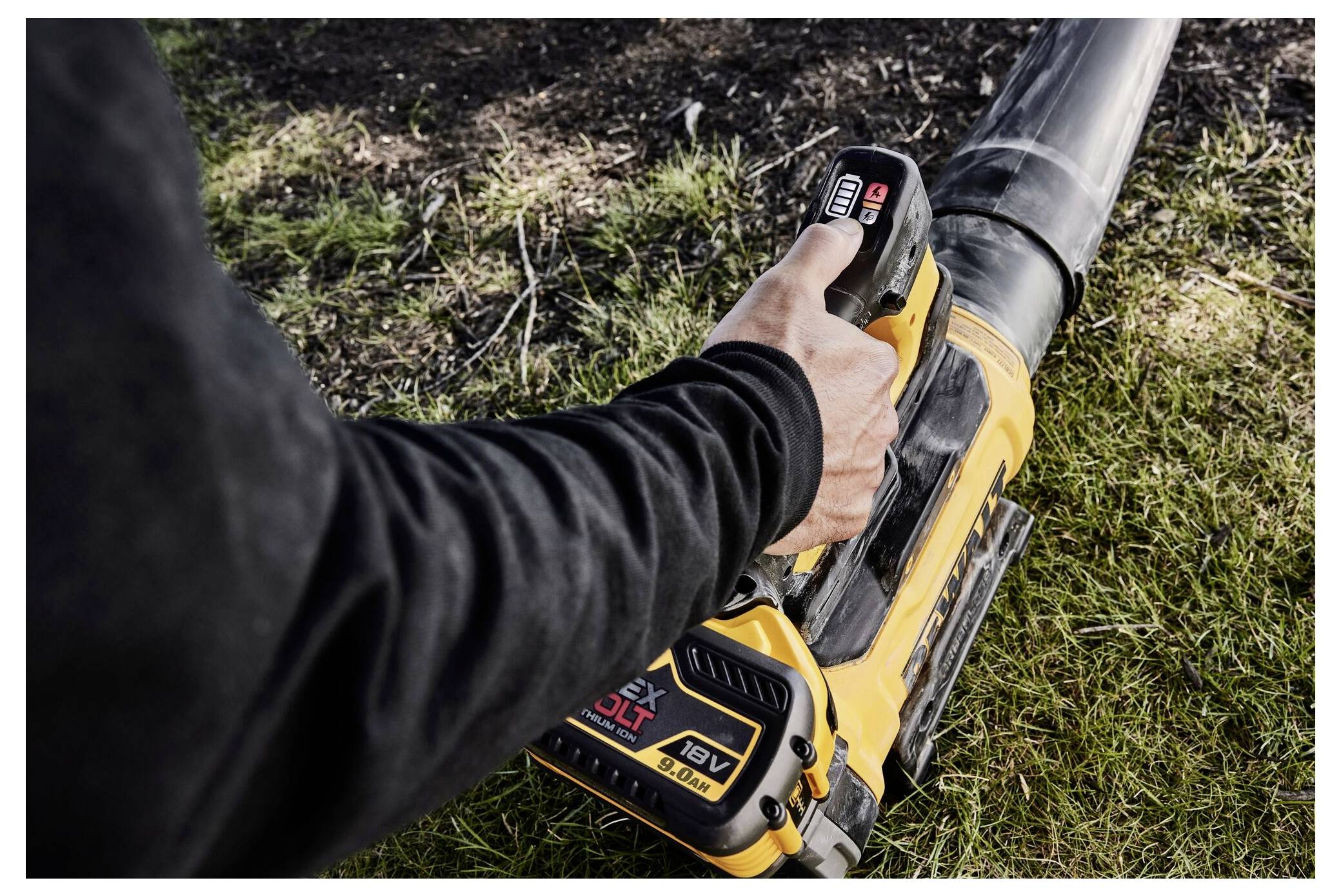 A person operates a yellow cordless leaf blower on grass, holding it with one hand. The blower is powered by an 18V battery.