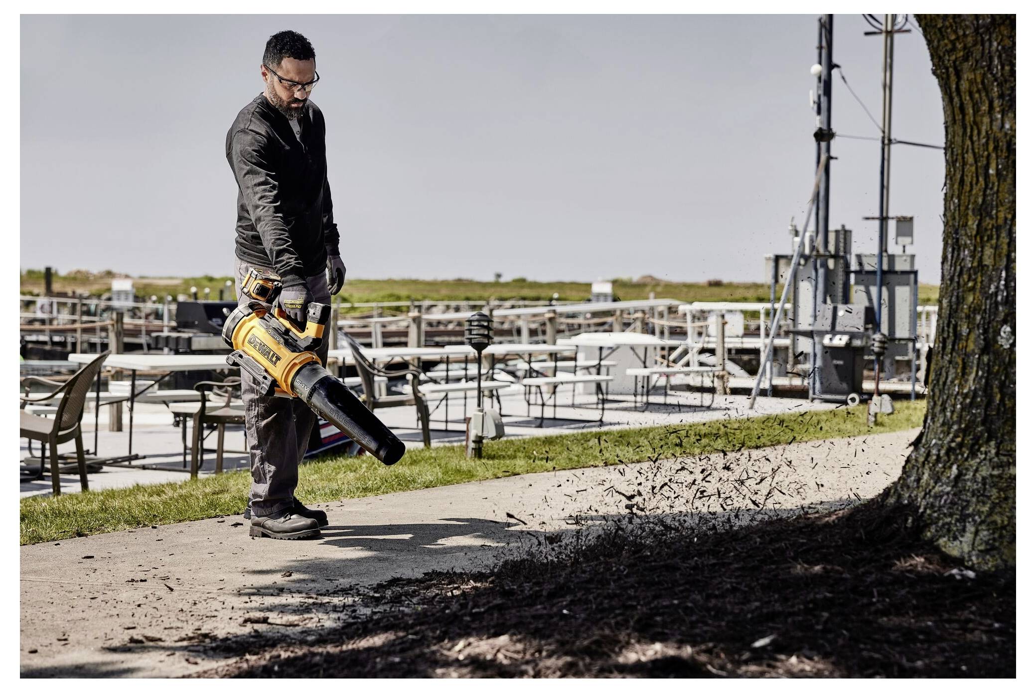 Man using a leaf blower to clear debris from a path near a waterfront with tables and chairs, cloudy sky in the background.