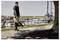 Man using a leaf blower to clear debris from a path near a waterfront with tables and chairs, cloudy sky in the background.