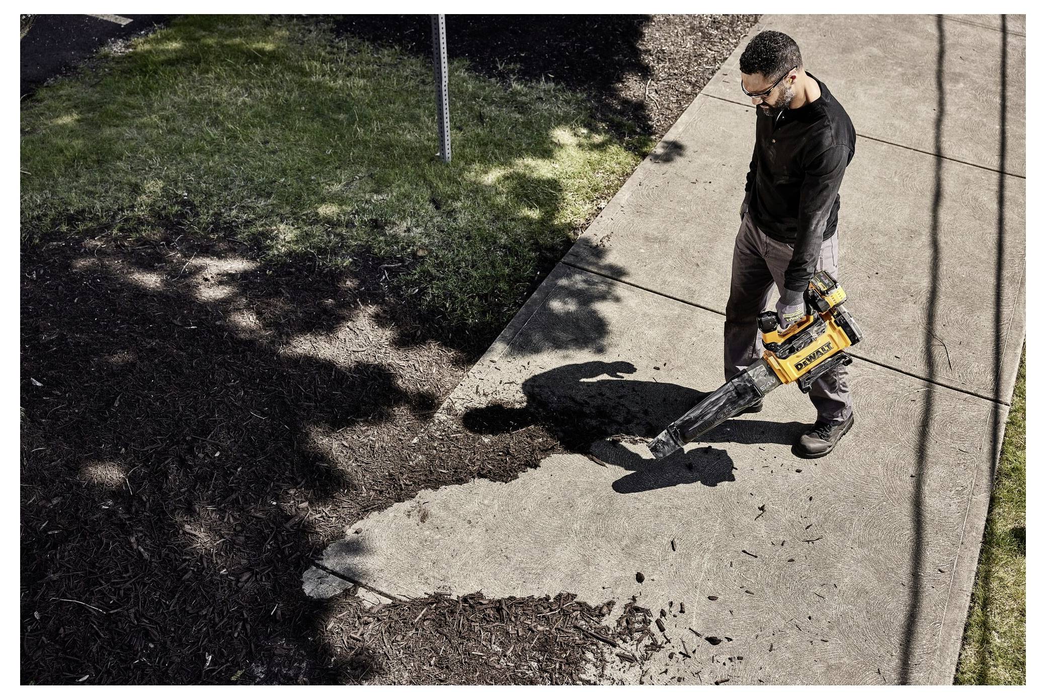 A person using a leaf blower clears debris from a paved walkway beside a garden area, under bright daylight and partial shadow.