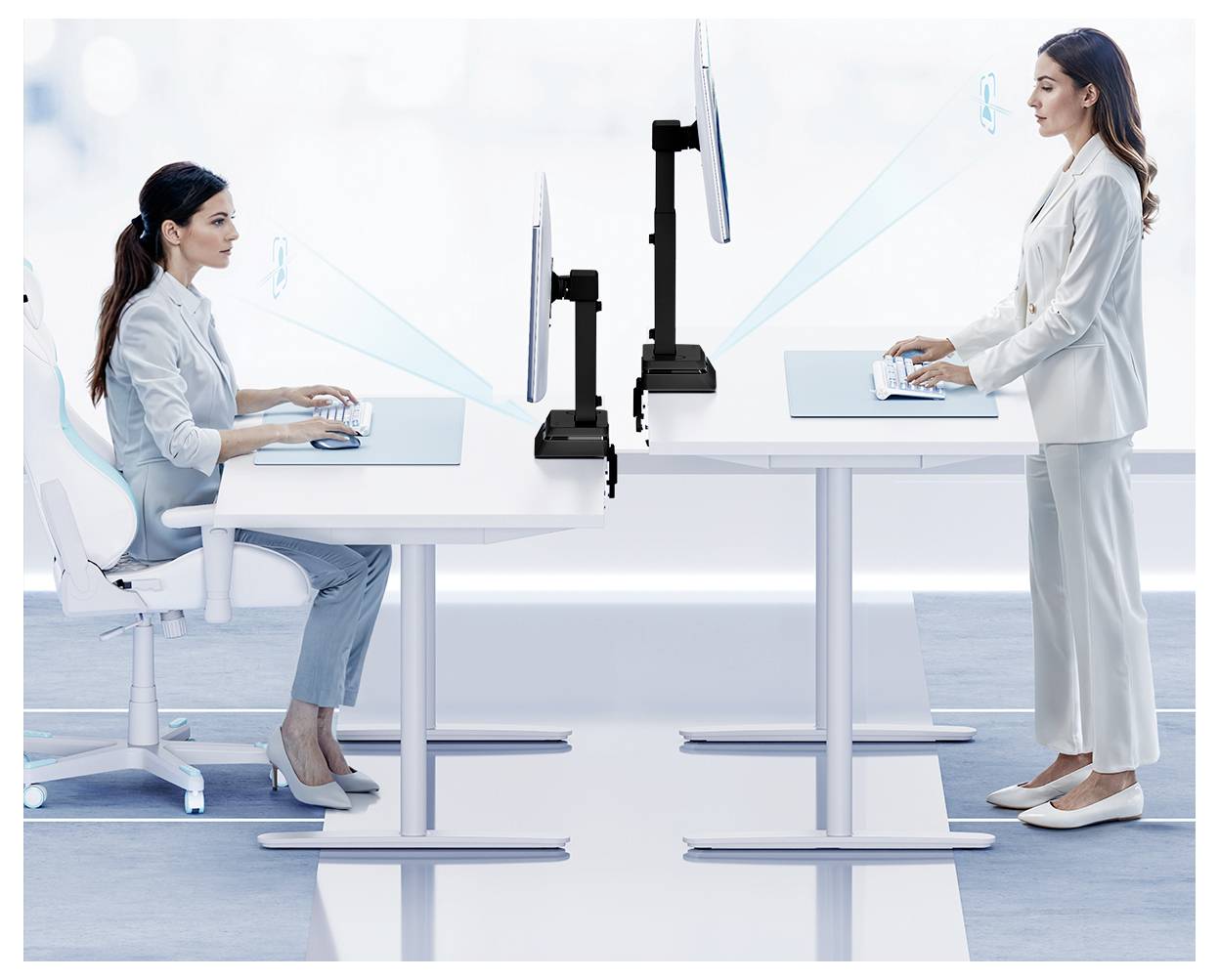 Two women in a modern office setting work at adjustable desks facing each other, one seated and the other standing, both using computers.